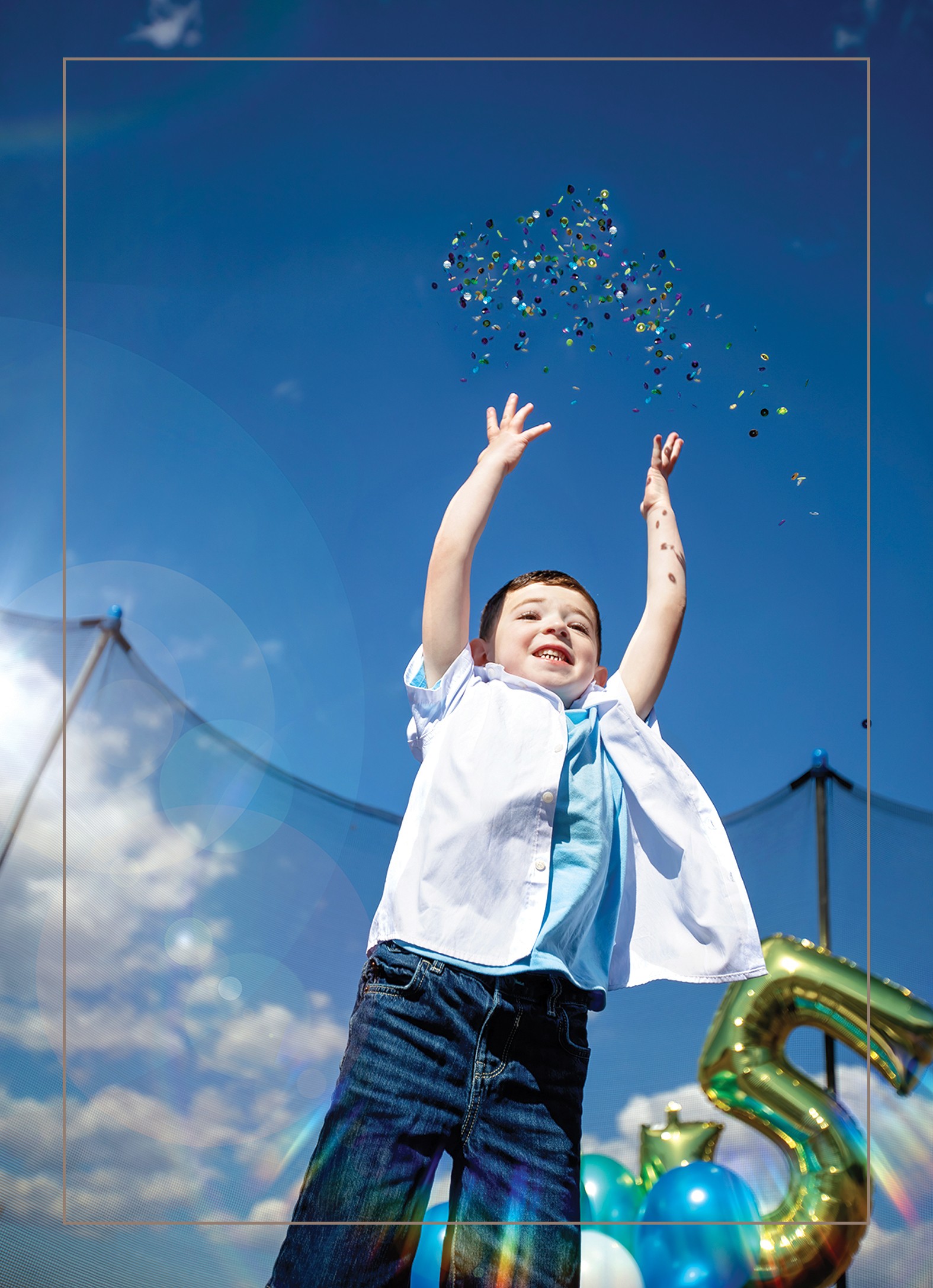 Child with arms raised, smiling, outdoors with blue sky and clouds, celebrating near large gold and blue balloons, including a number five.