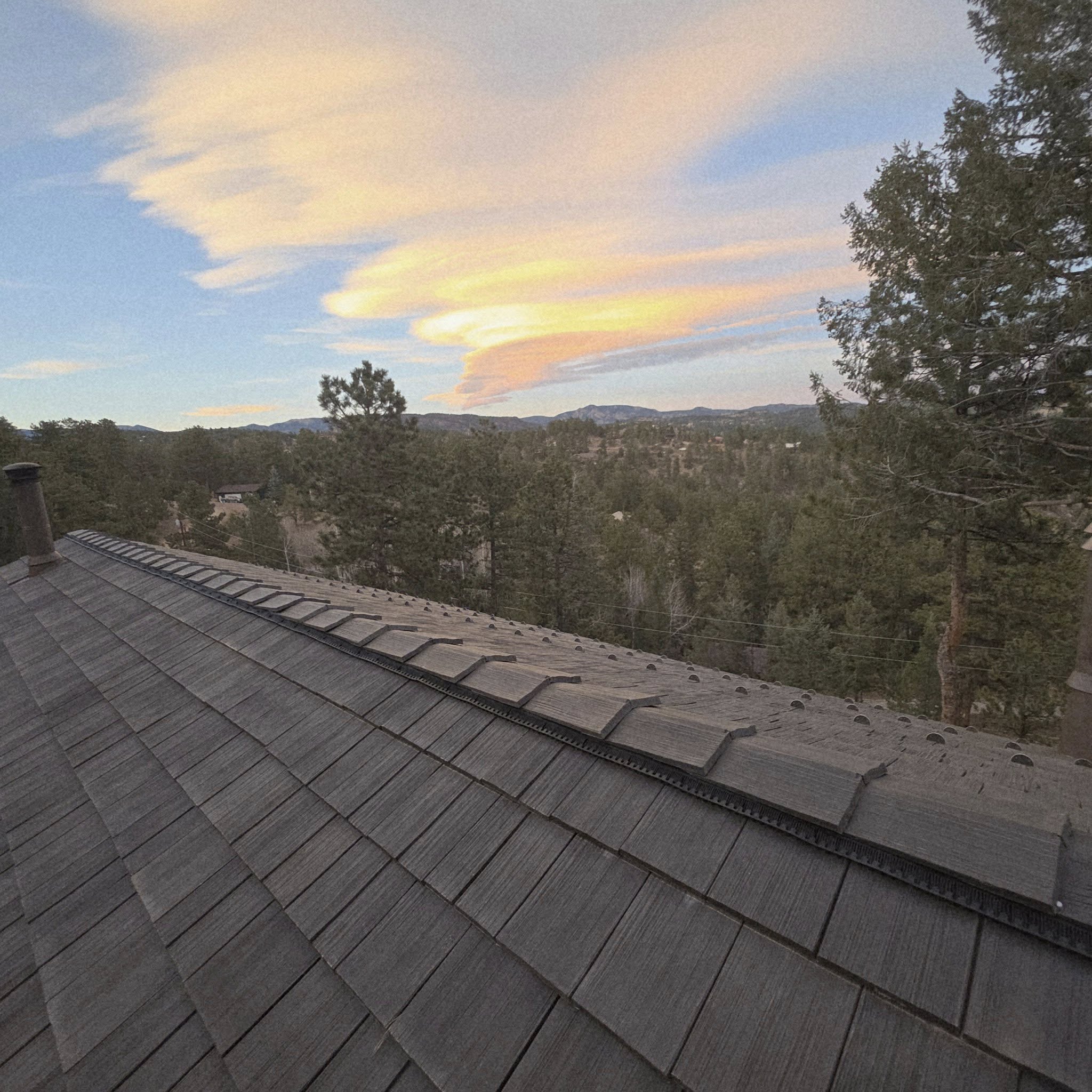 View of a roof with shingles, trees, and a sky with clouds and sunset colors.