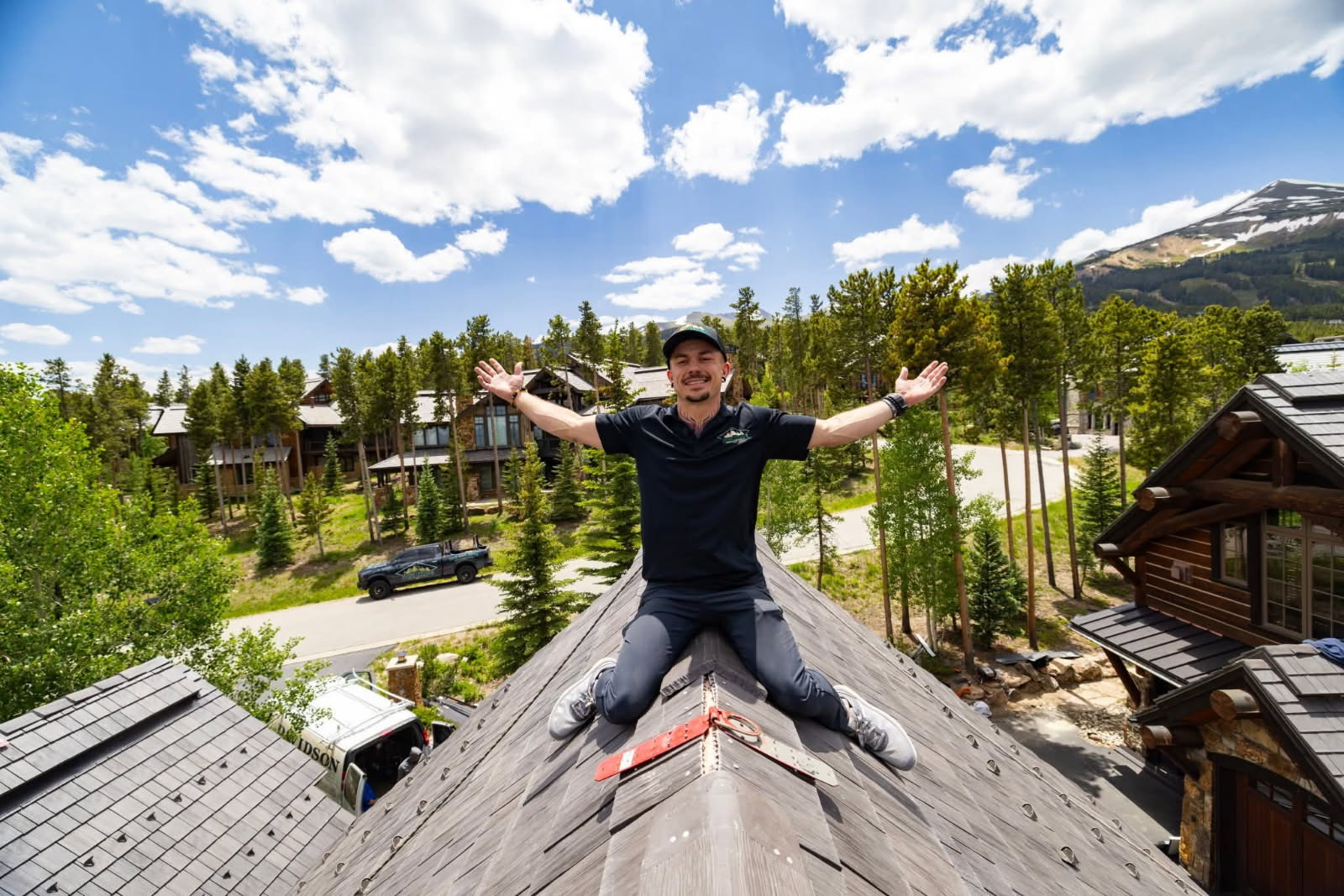 Person sitting on a rooftop with arms outstretched, surrounded by trees and buildings, under a partly cloudy sky.