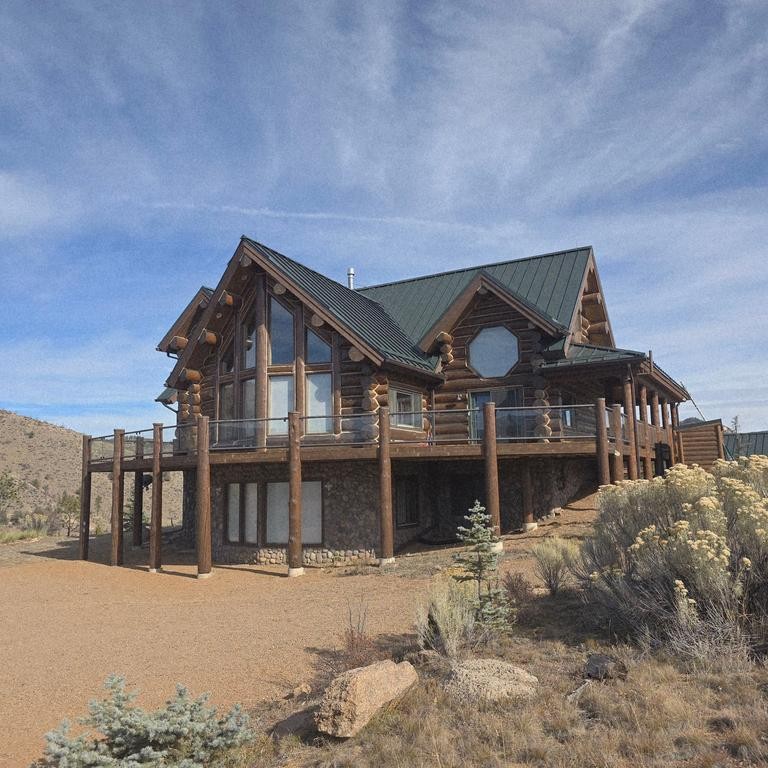 Large wooden house with a green roof, elevated on stilts, surrounded by desert plants and rocks, under a blue sky.
