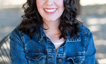 Young woman with dark wavy hair smiling outdoors, wearing a denim jacket, arms crossed, trees in background.