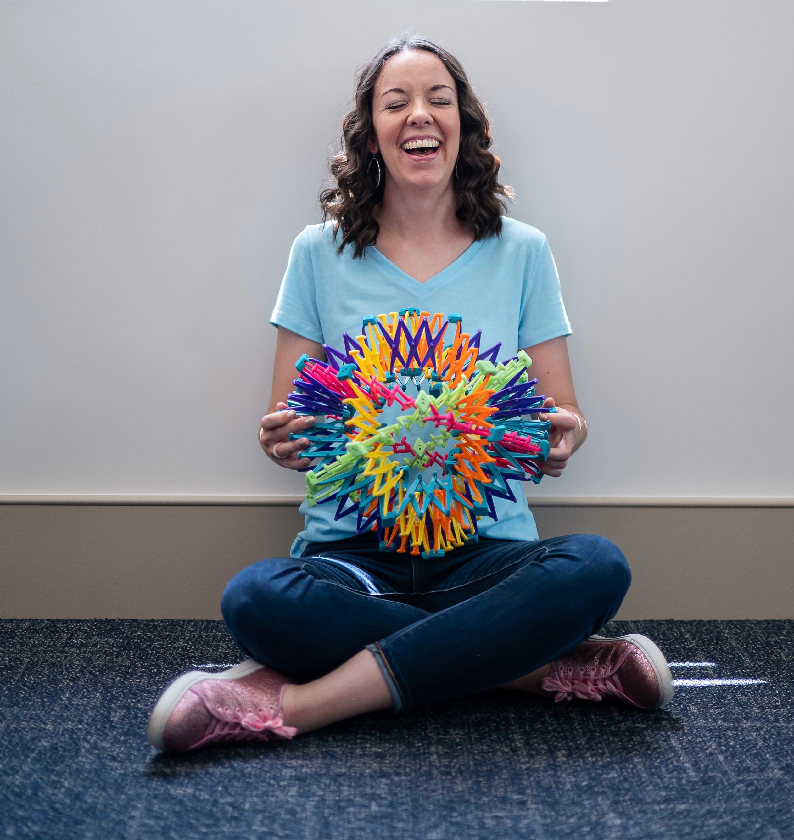 Woman sitting cross-legged on floor, holding colorful spiky ball, smiling, against plain wall background.