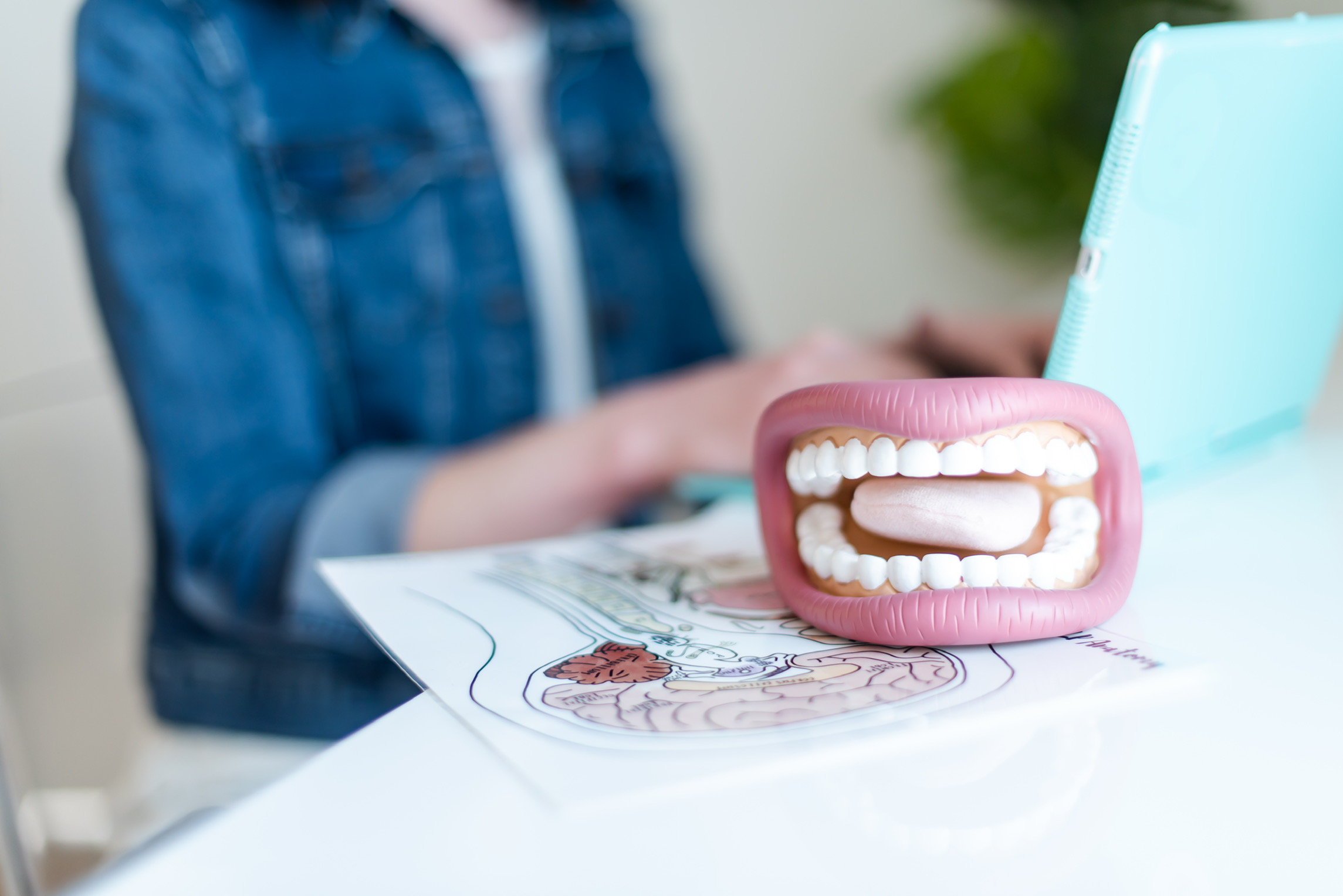 A pink toy with a mouth and teeth is on a table, with a person using a laptop in the background.