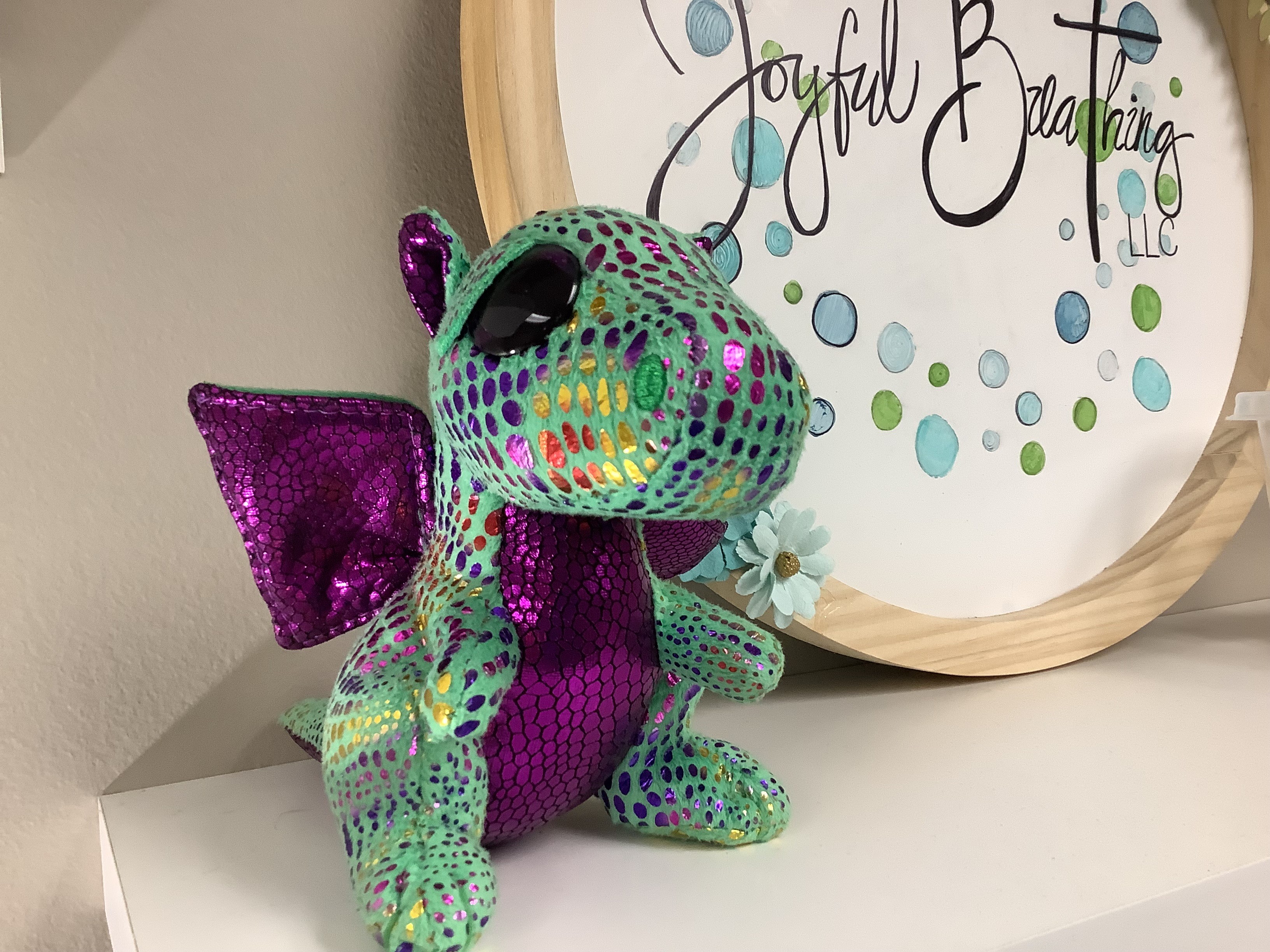 Colorful plush dragon with purple wings and a purple belly, sitting on a white surface next to a decorative sign.