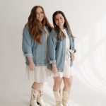 Two young women stand side by side, smiling, wearing denim jackets, white dresses, and cowboy boots, against a plain white background.
