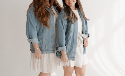 Two young women stand side by side, smiling, wearing denim jackets, white dresses, and cowboy boots, against a plain white background.