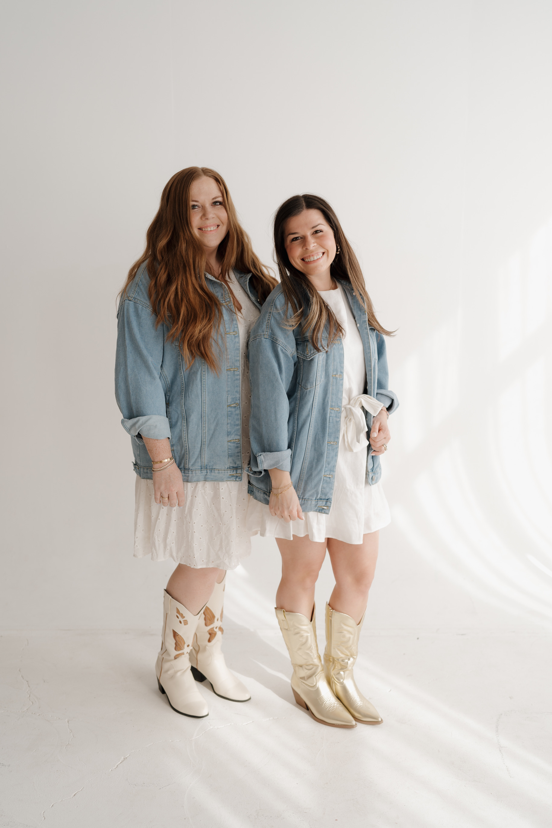 Two young women stand side by side, smiling, wearing denim jackets, white dresses, and cowboy boots, against a plain white background.