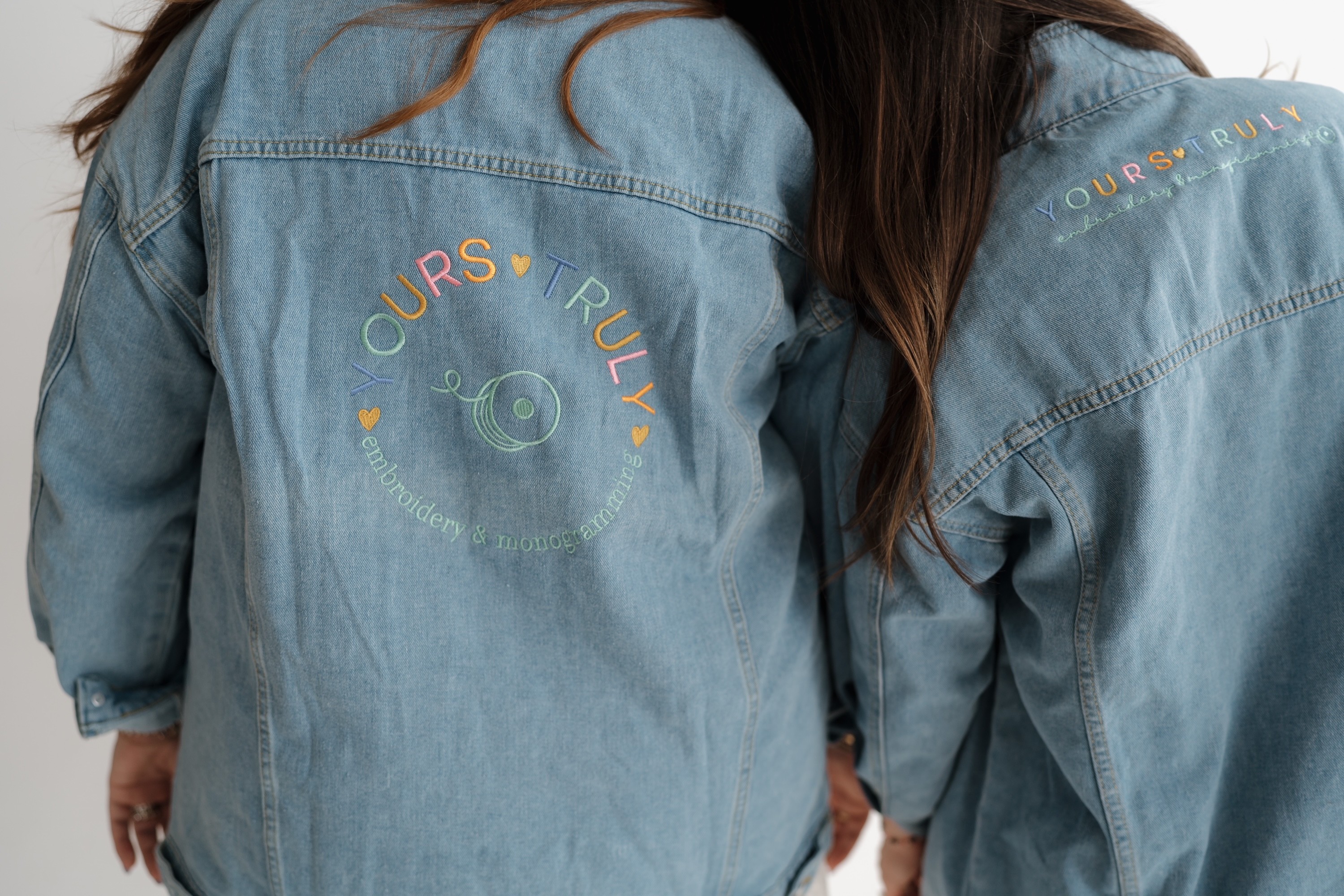 Two people wearing denim jackets with embroidered text and designs on the back, standing close together.
