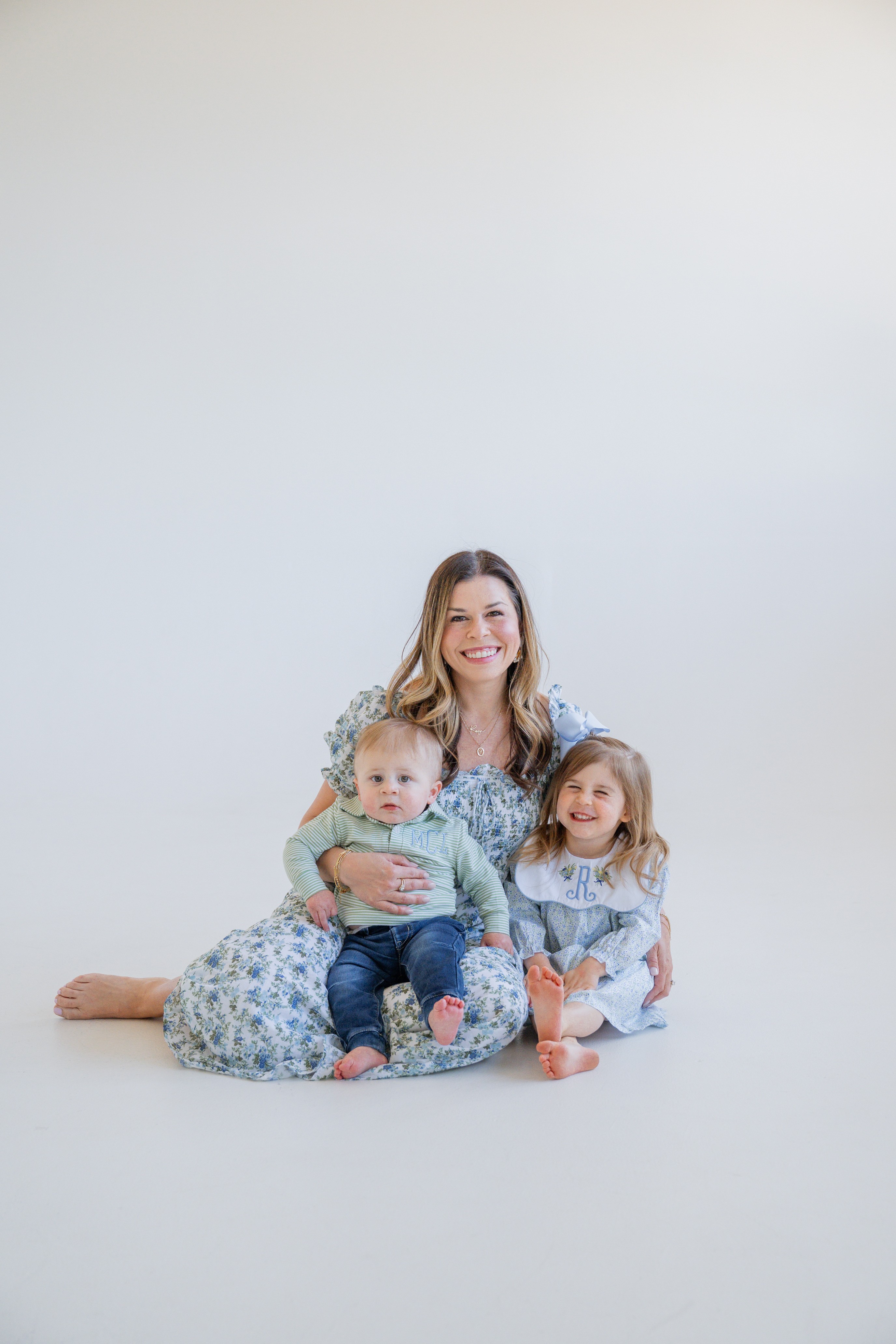 Woman sitting on the floor with two children, all smiling, against a plain light background.