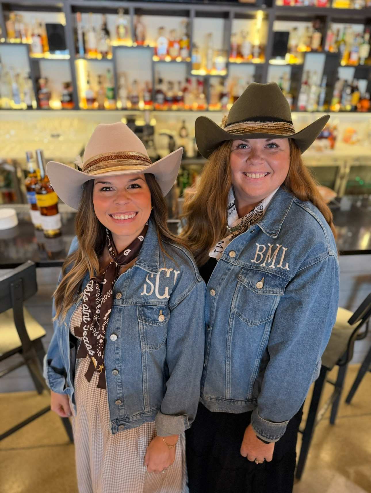 Two women wearing cowboy hats and denim jackets smiling in a bar or restaurant setting.