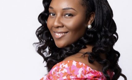 Smiling woman with curly hair wearing a pink floral off-shoulder top, facing slightly to the right against a white background.