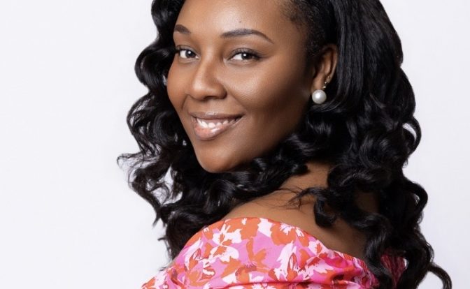 Smiling woman with curly hair wearing a pink floral off-shoulder top, facing slightly to the right against a white background.