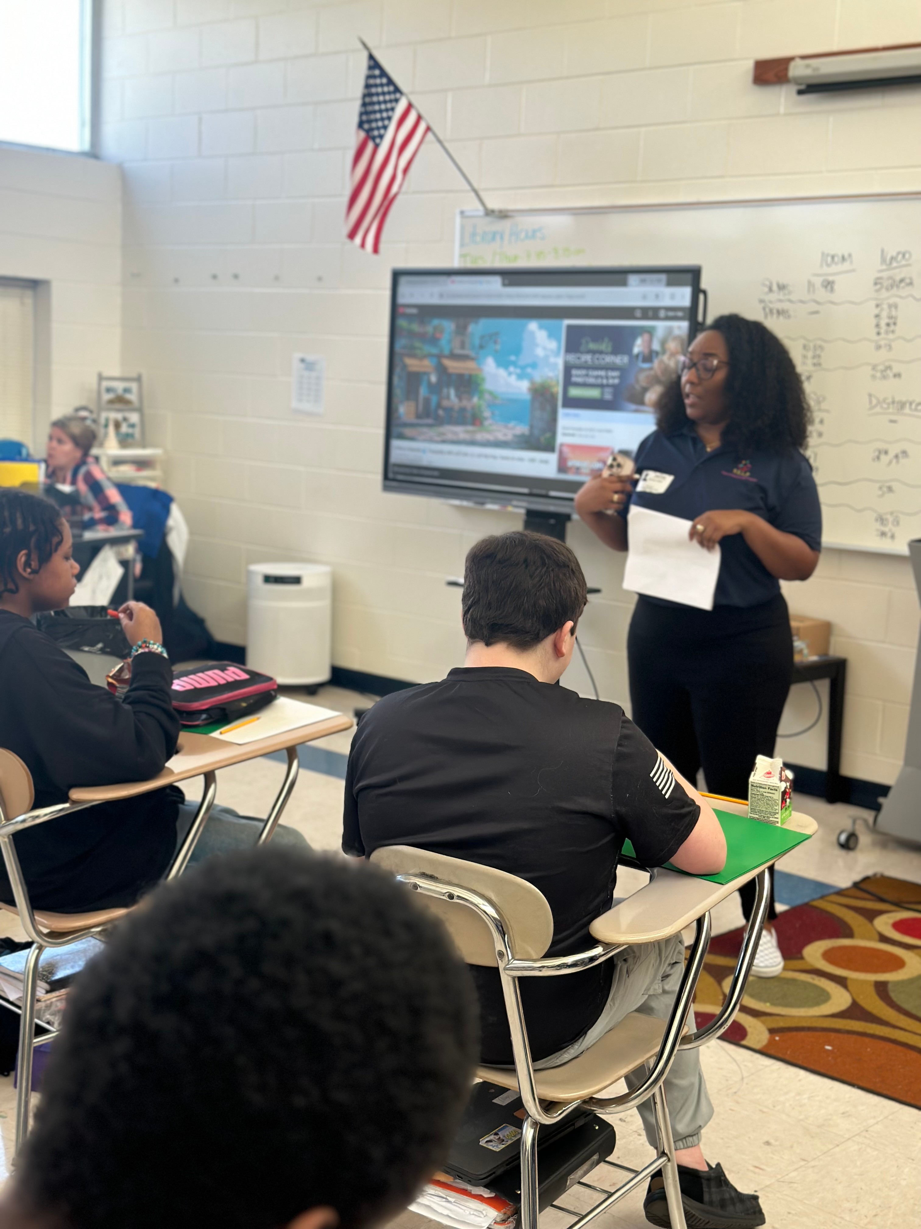Teacher standing in front of a classroom with students seated at desks, a large screen behind her displays a presentation, American flag on wall.