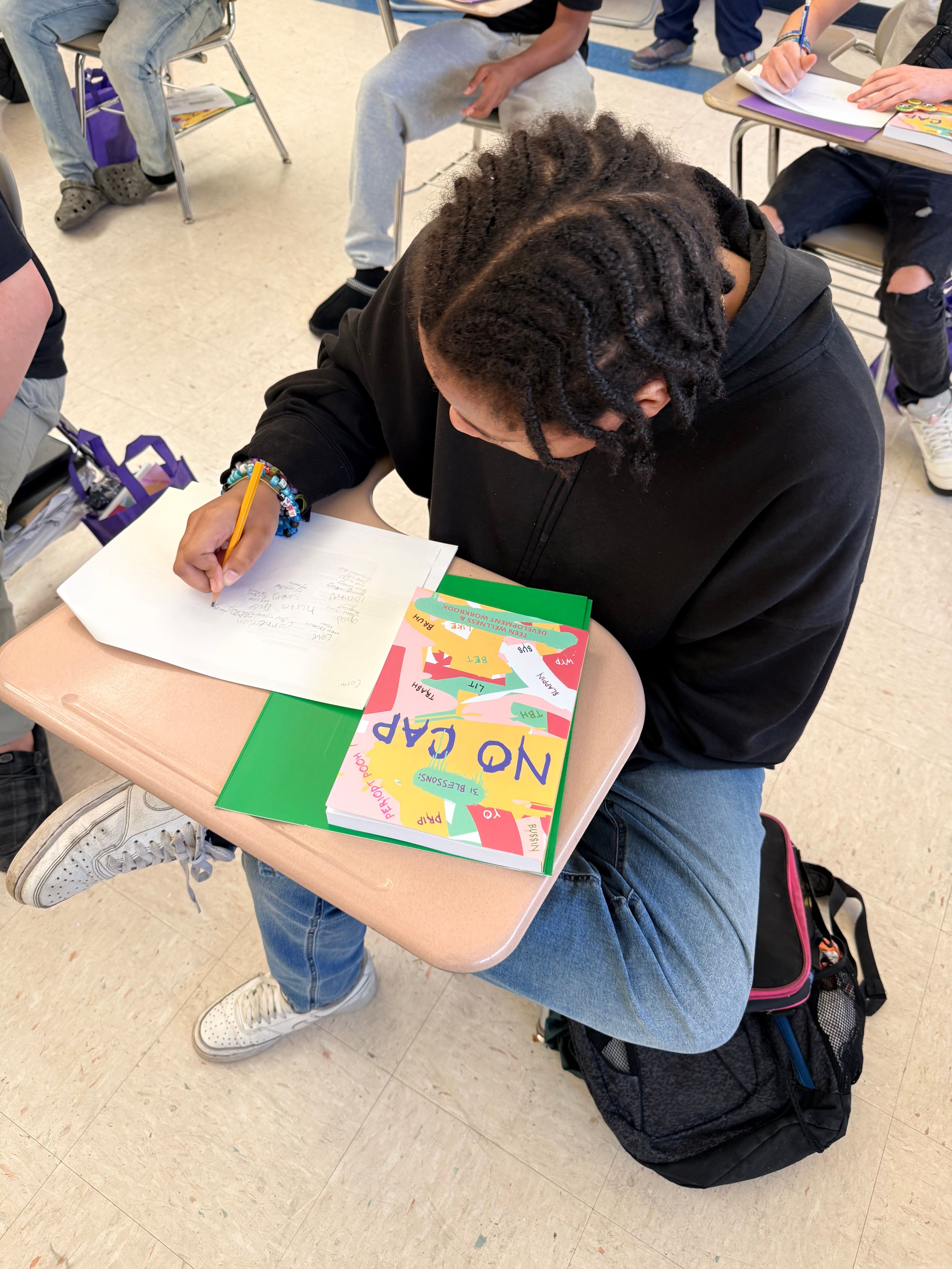 Person with dreadlocks writing on paper at desk with colorful notebook and backpack nearby.