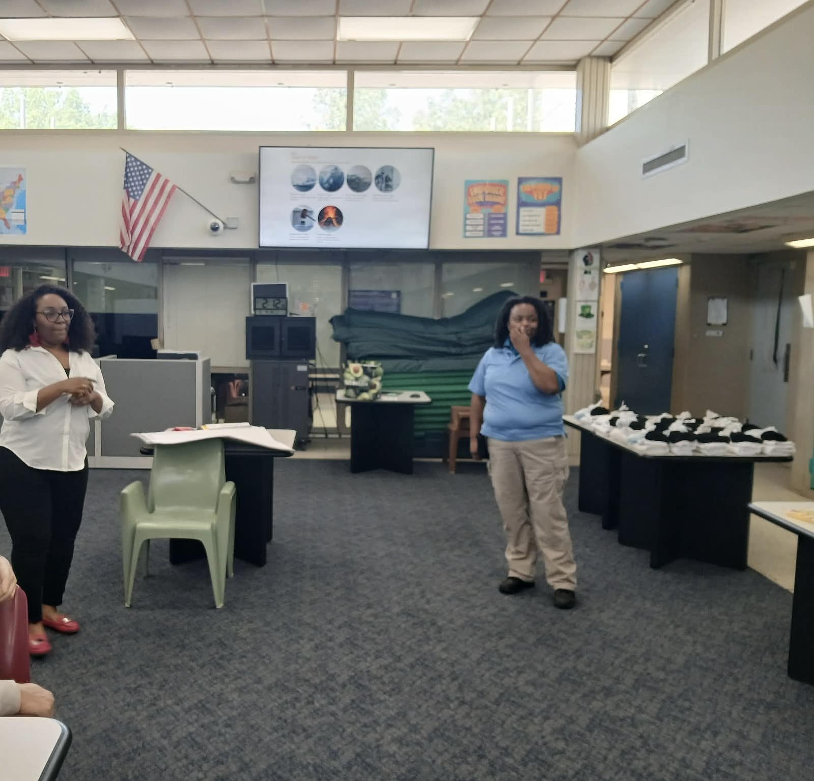 Two women standing in a classroom with tables, chairs, and educational posters on the wall.