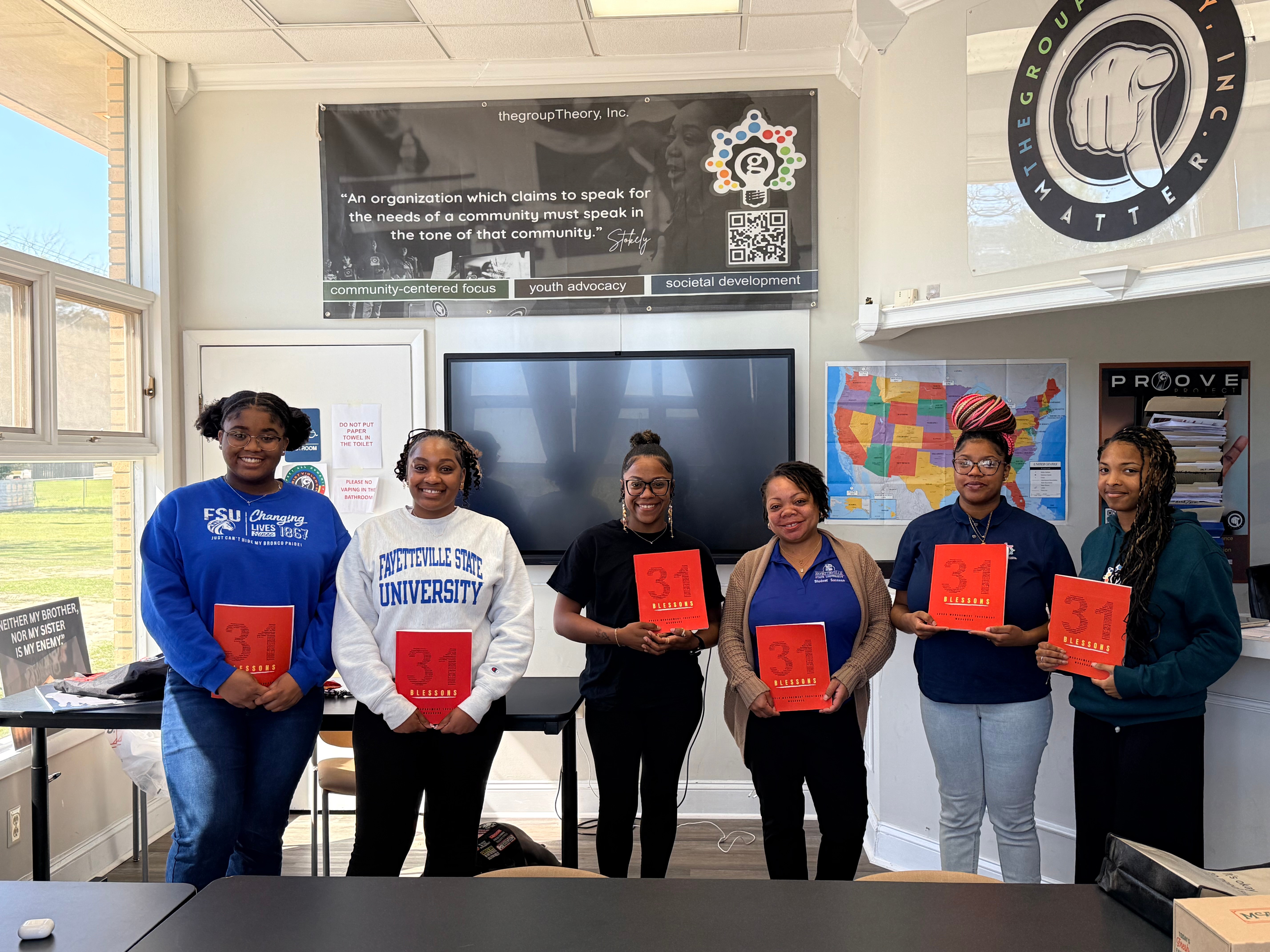 Six women standing in a classroom holding red certificates, smiling, with a large screen and educational posters behind them.