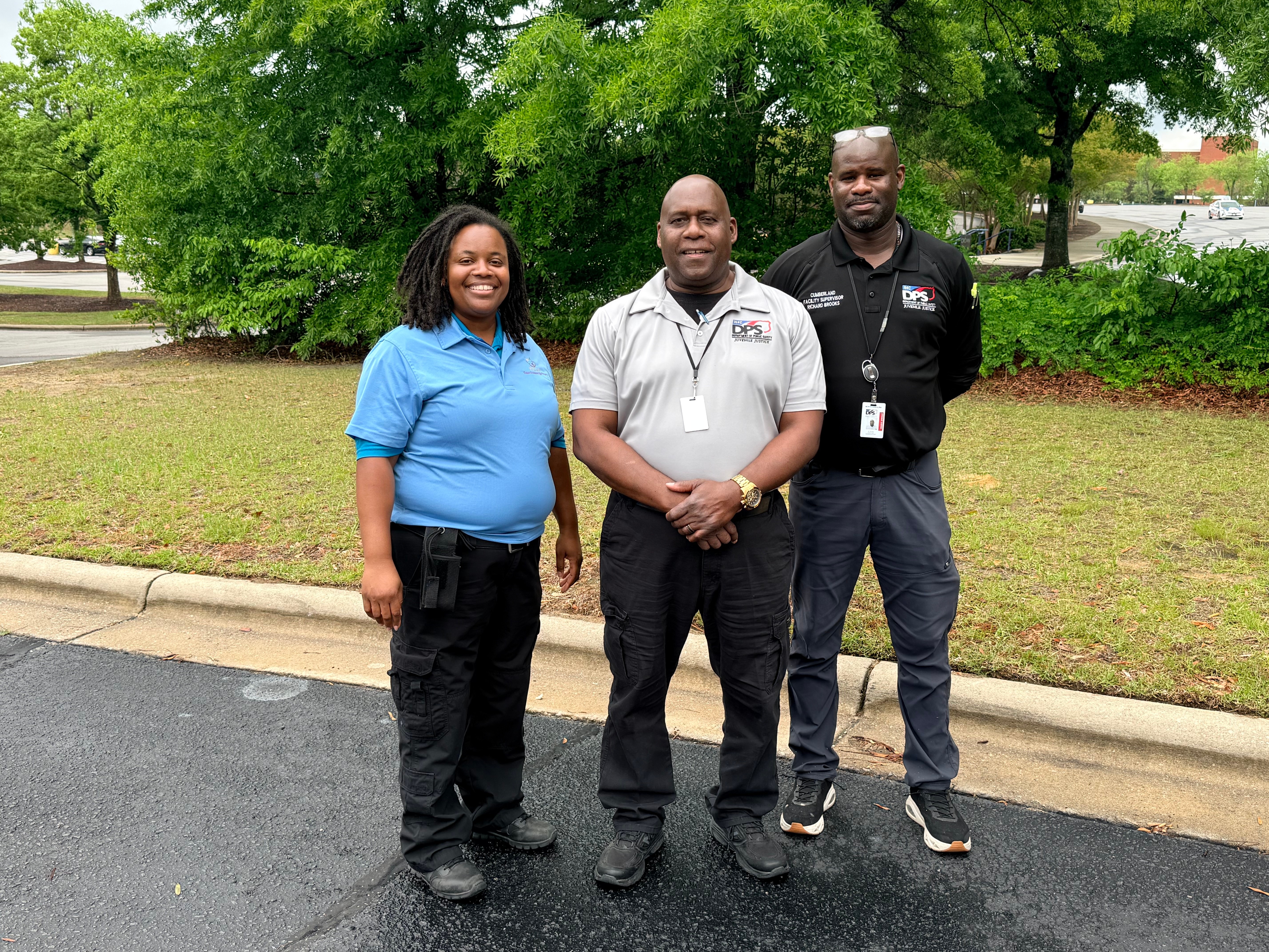 Three people standing outdoors on a paved area with trees and grass behind them, smiling at the camera.
