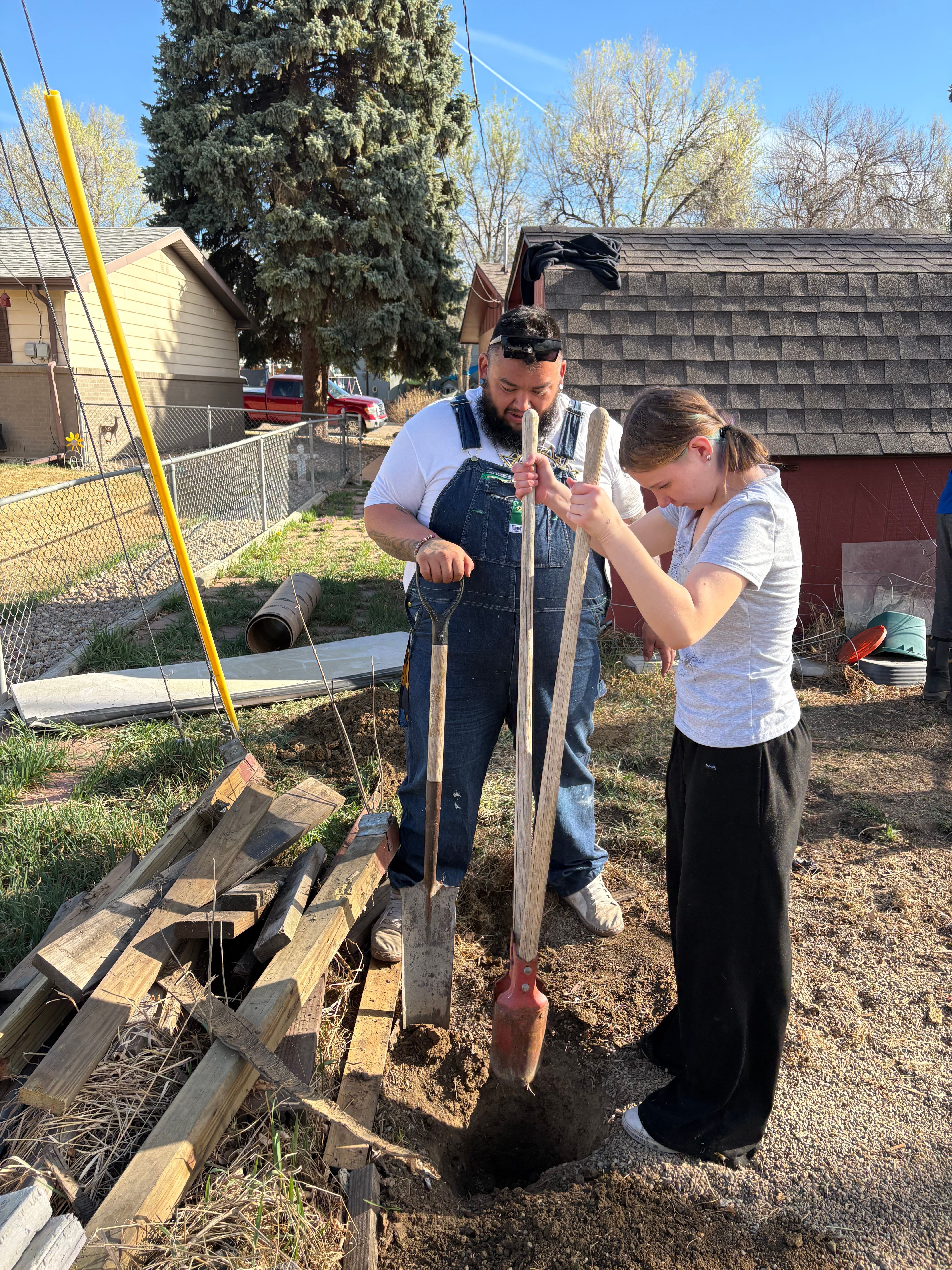 Two women working together on a construction project outdoors, with wood planks and tools nearby, under a clear sky.