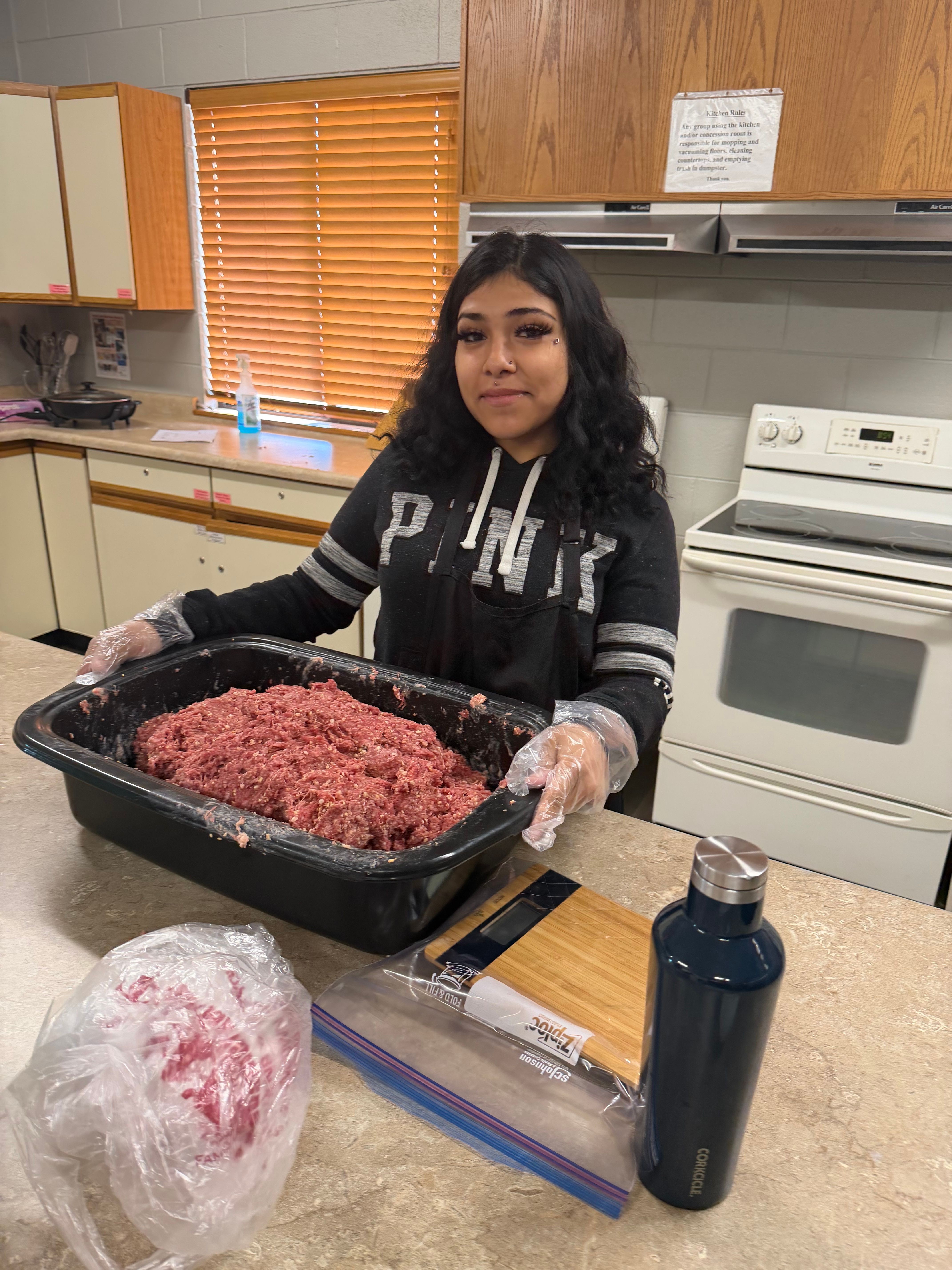 Woman wearing gloves handling raw ground meat on a black tray in a kitchen. Various kitchen items are visible.