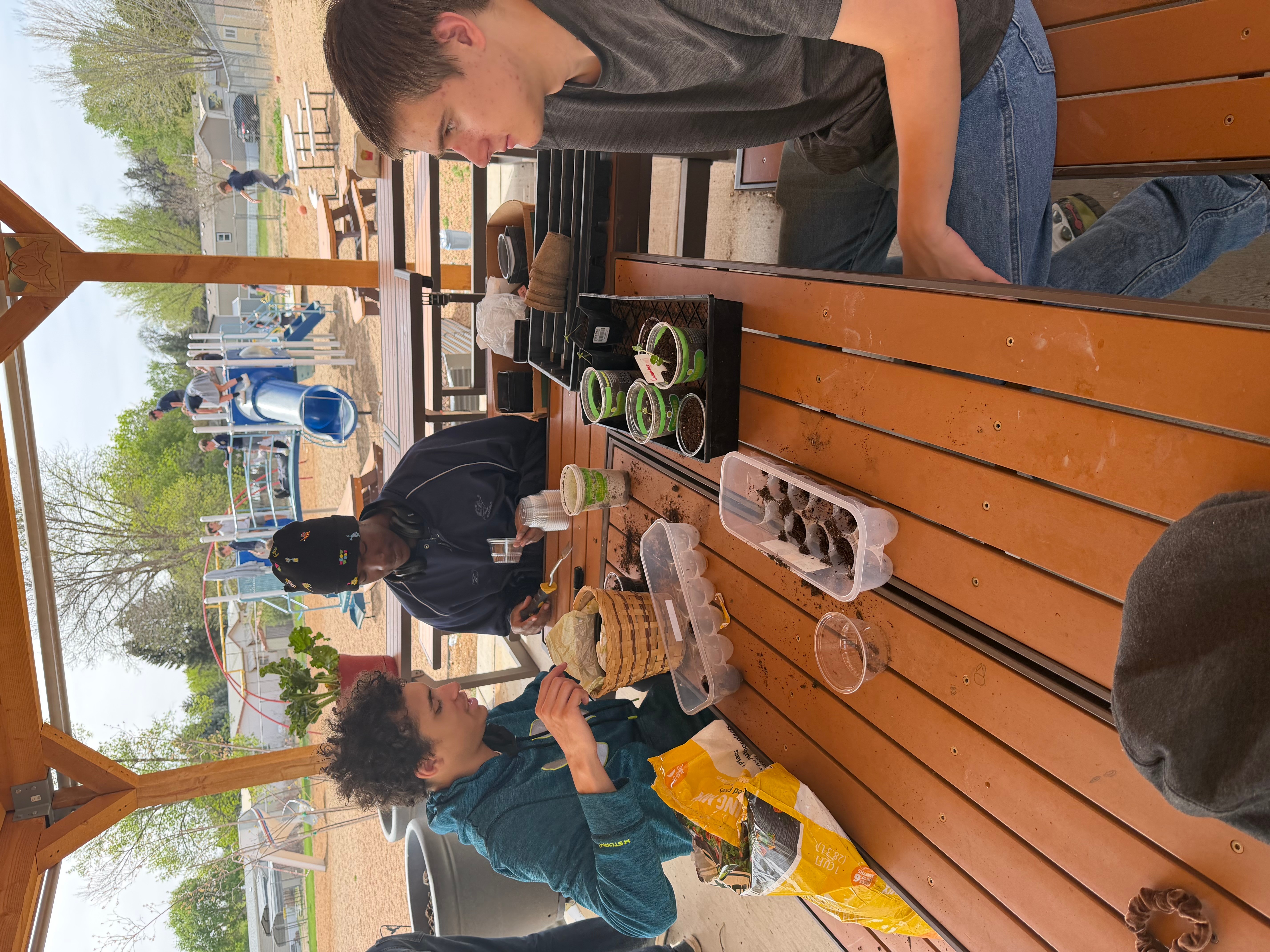 Two children sit at a wooden table outdoors, eating food and drinking from bottles, with other people and playground equipment in background.