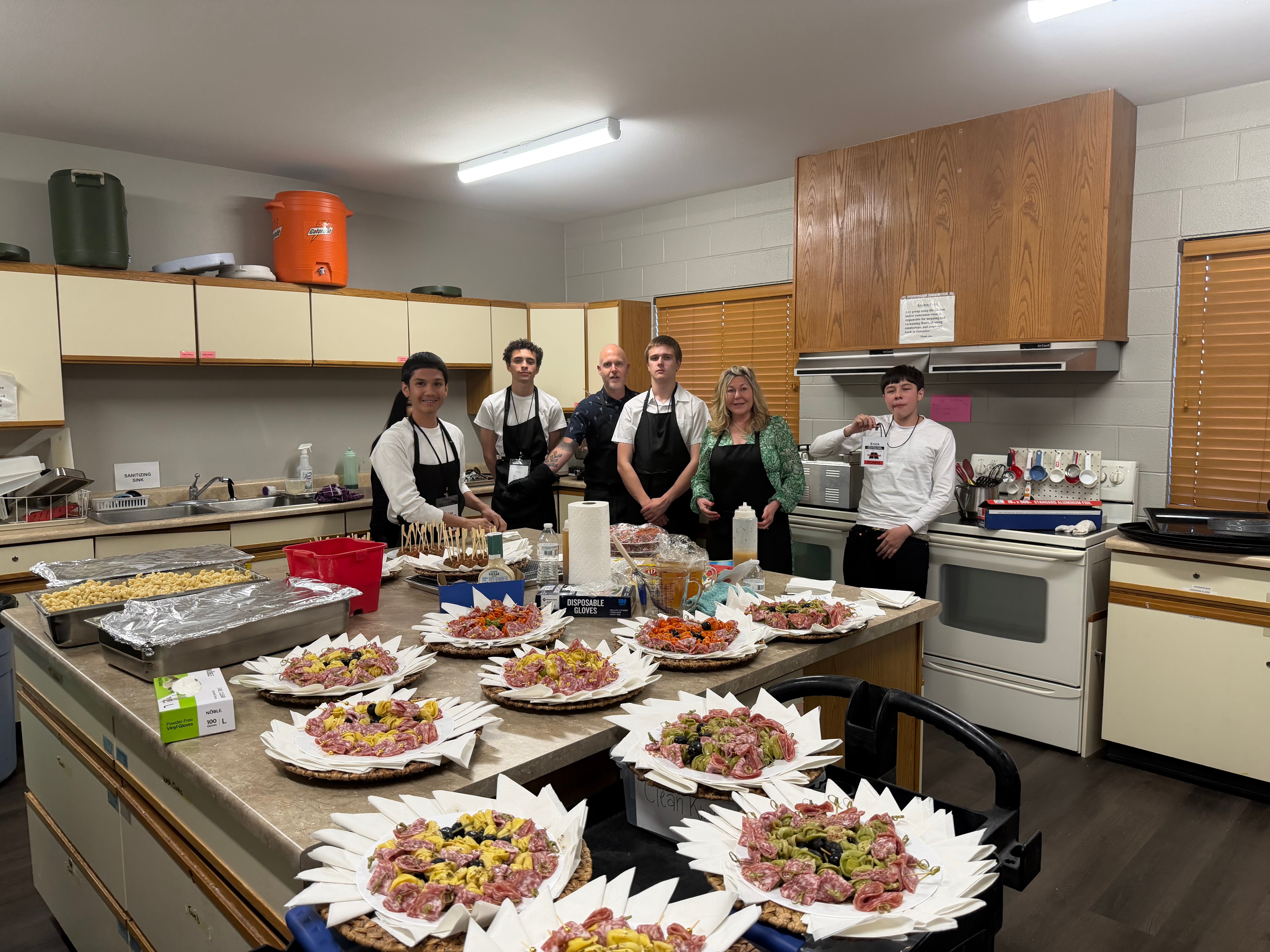 Group of six people standing behind a table with food in a kitchen, preparing for a meal or event.