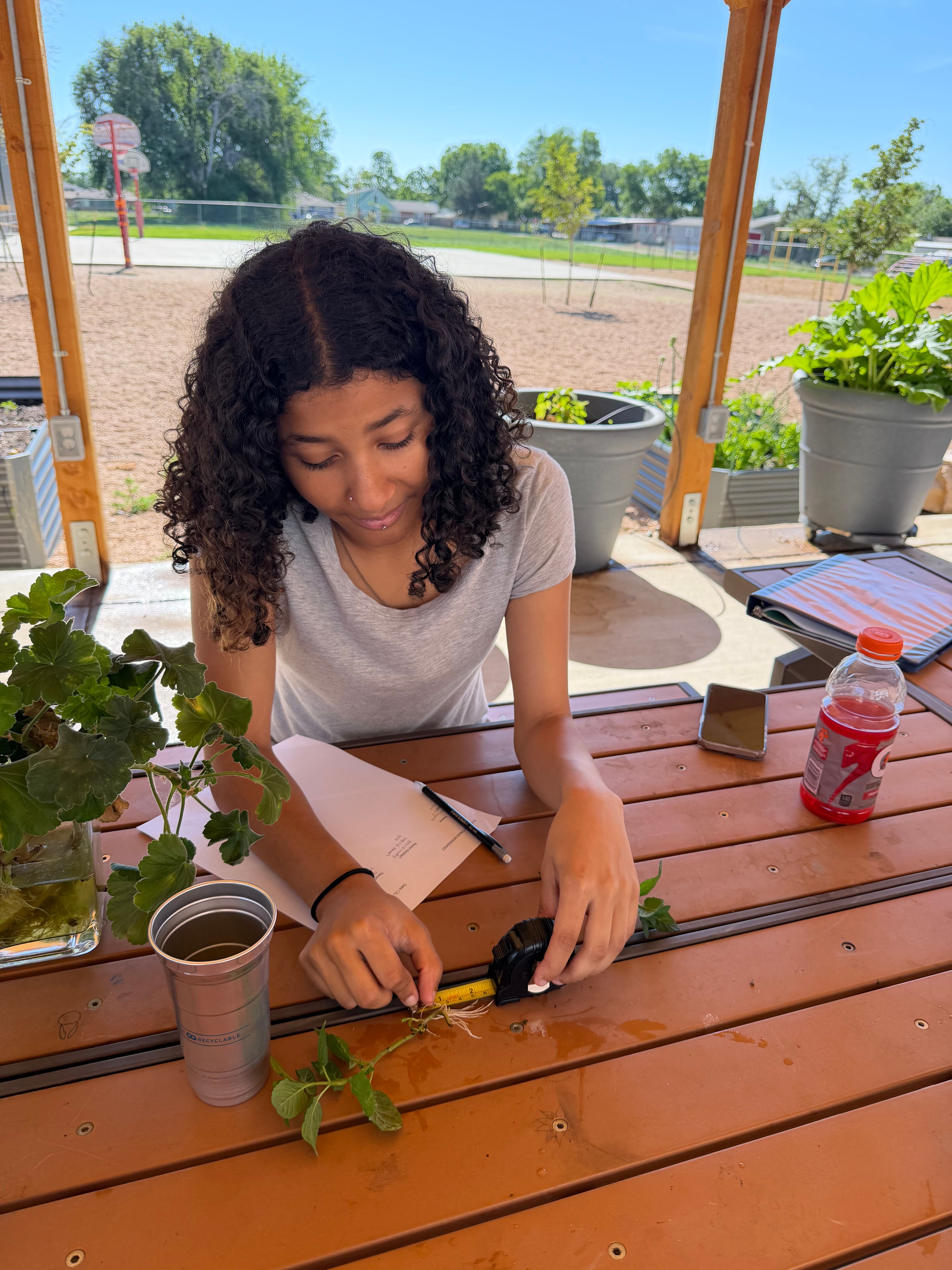 Person with curly hair writing on paper at outdoor wooden table with plants and a drink, sunny day, trees in background.