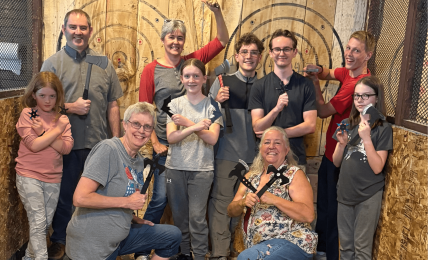 Group of ten people, some holding axes, posing indoors with wooden targets and banners in the background.