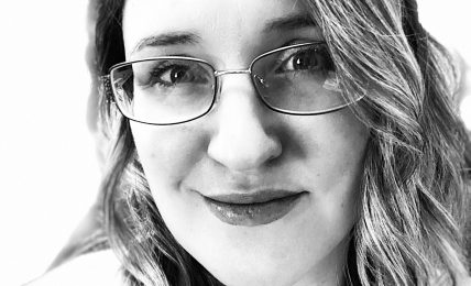 Woman with glasses and wavy hair smiling, looking at camera, in black and white.