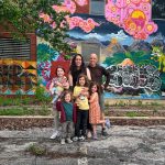 Group of six people, including children and adults, standing in front of a colorful mural with trees and graffiti.