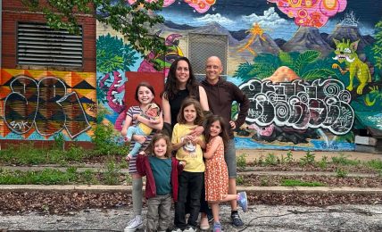 Group of six people, including children and adults, standing in front of a colorful mural with trees and graffiti.