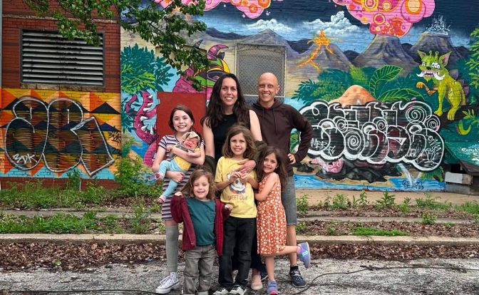 Group of six people, including children and adults, standing in front of a colorful mural with trees and graffiti.