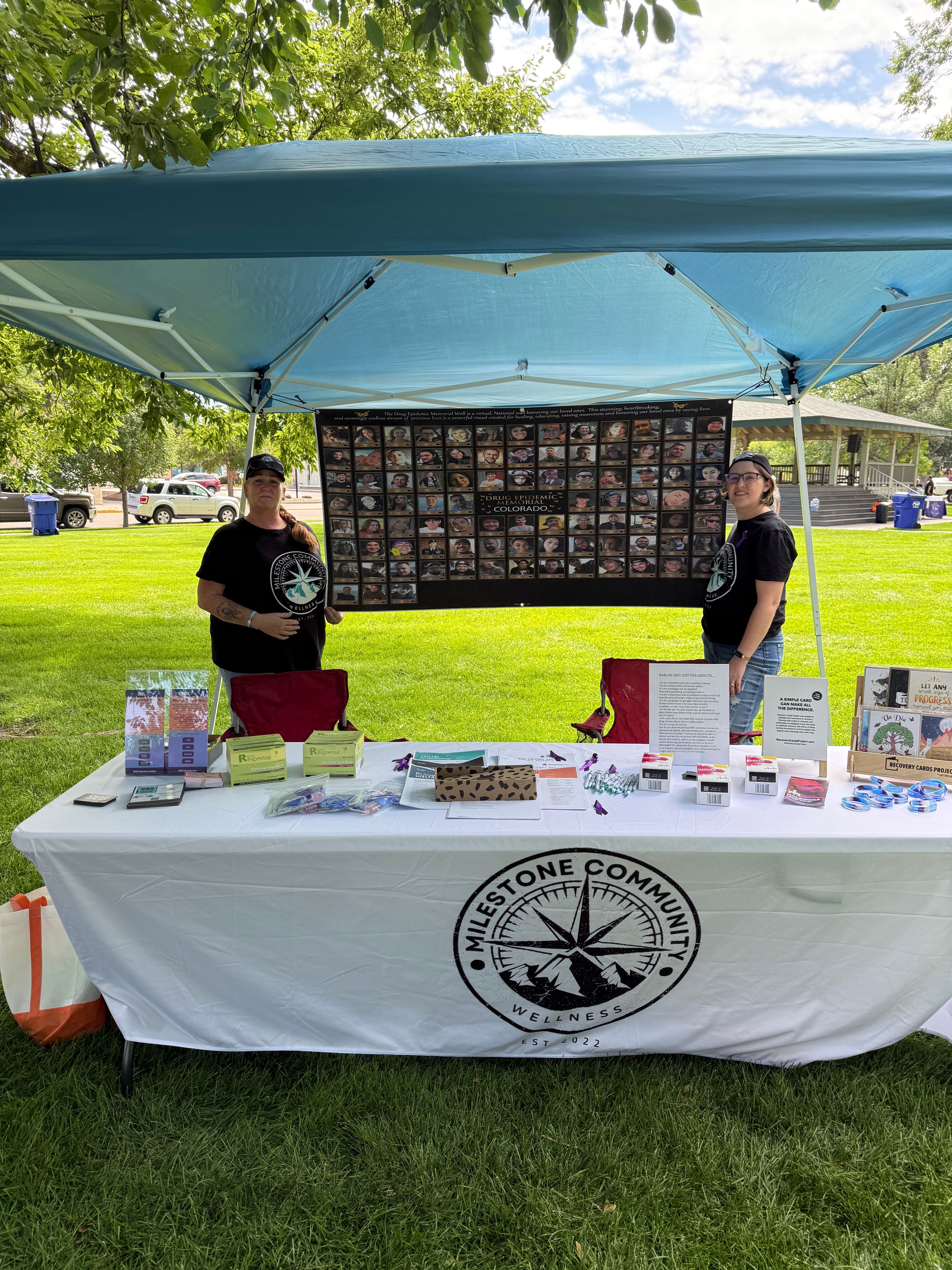 Two people at an outdoor community event with a table and display under a canopy, grass and trees in background.