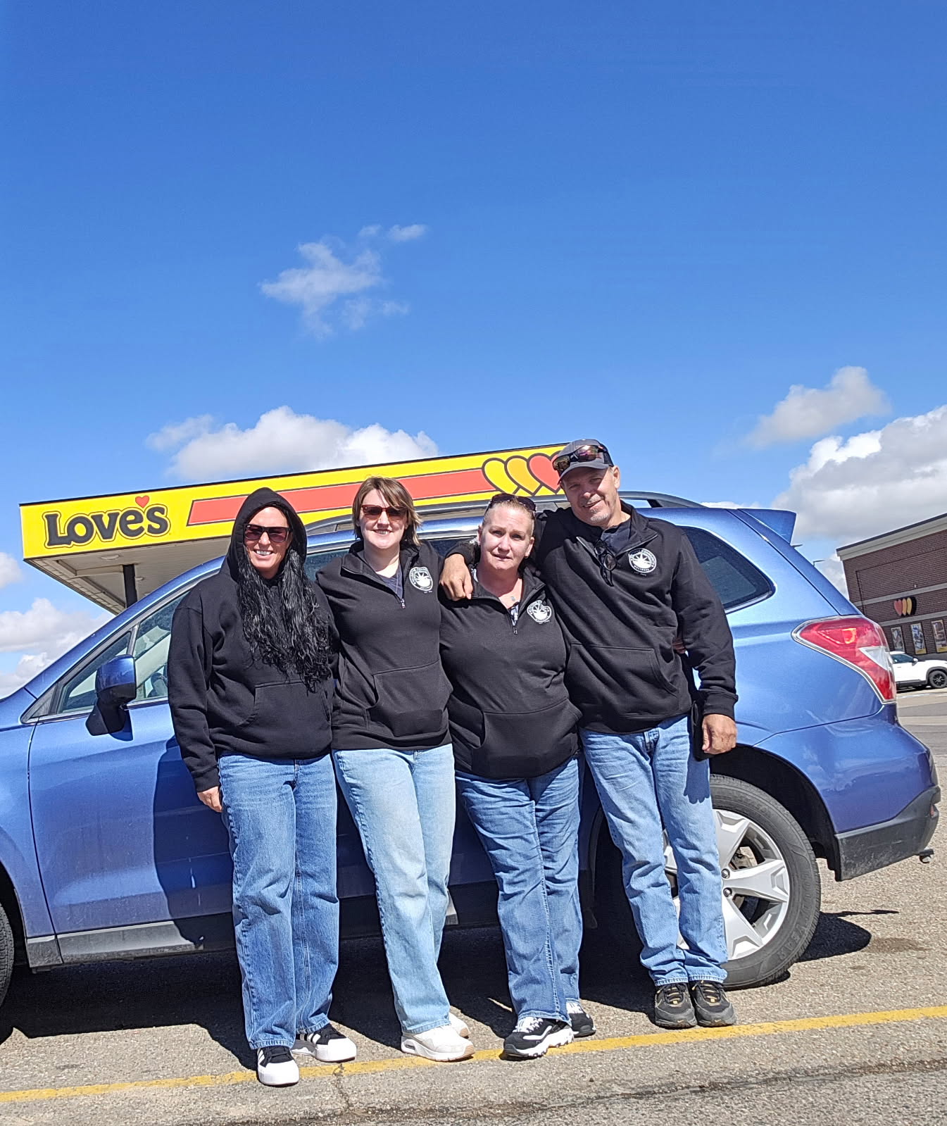 Four people stand in front of a blue car with a Love's sign, smiling outdoors on a sunny day.