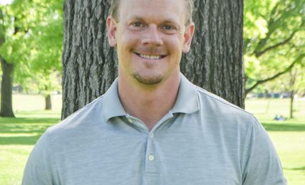 Man smiling outdoors in front of a tree with green leaves, wearing a light gray polo shirt.