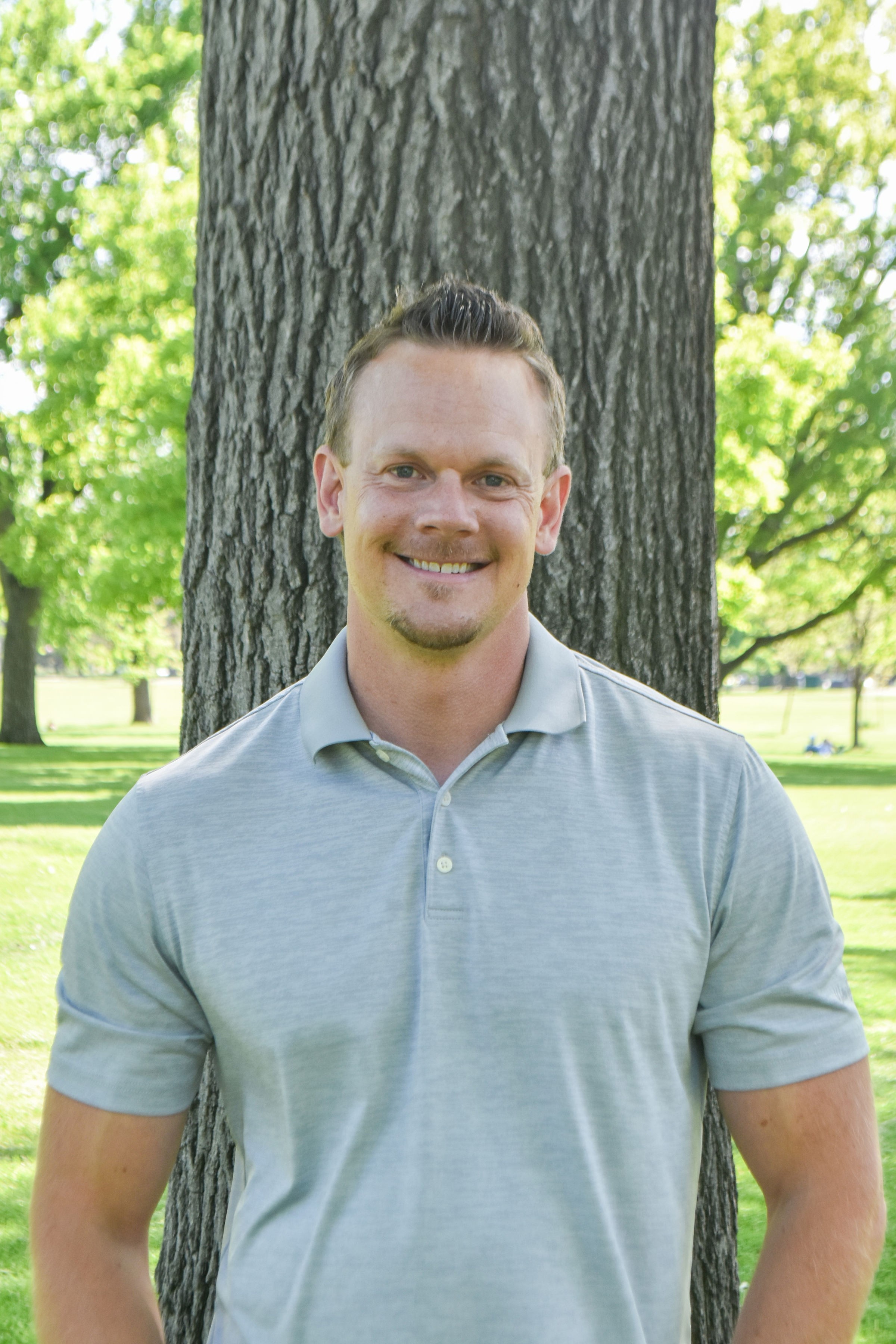 Man smiling outdoors in front of a tree with green leaves, wearing a light gray polo shirt.