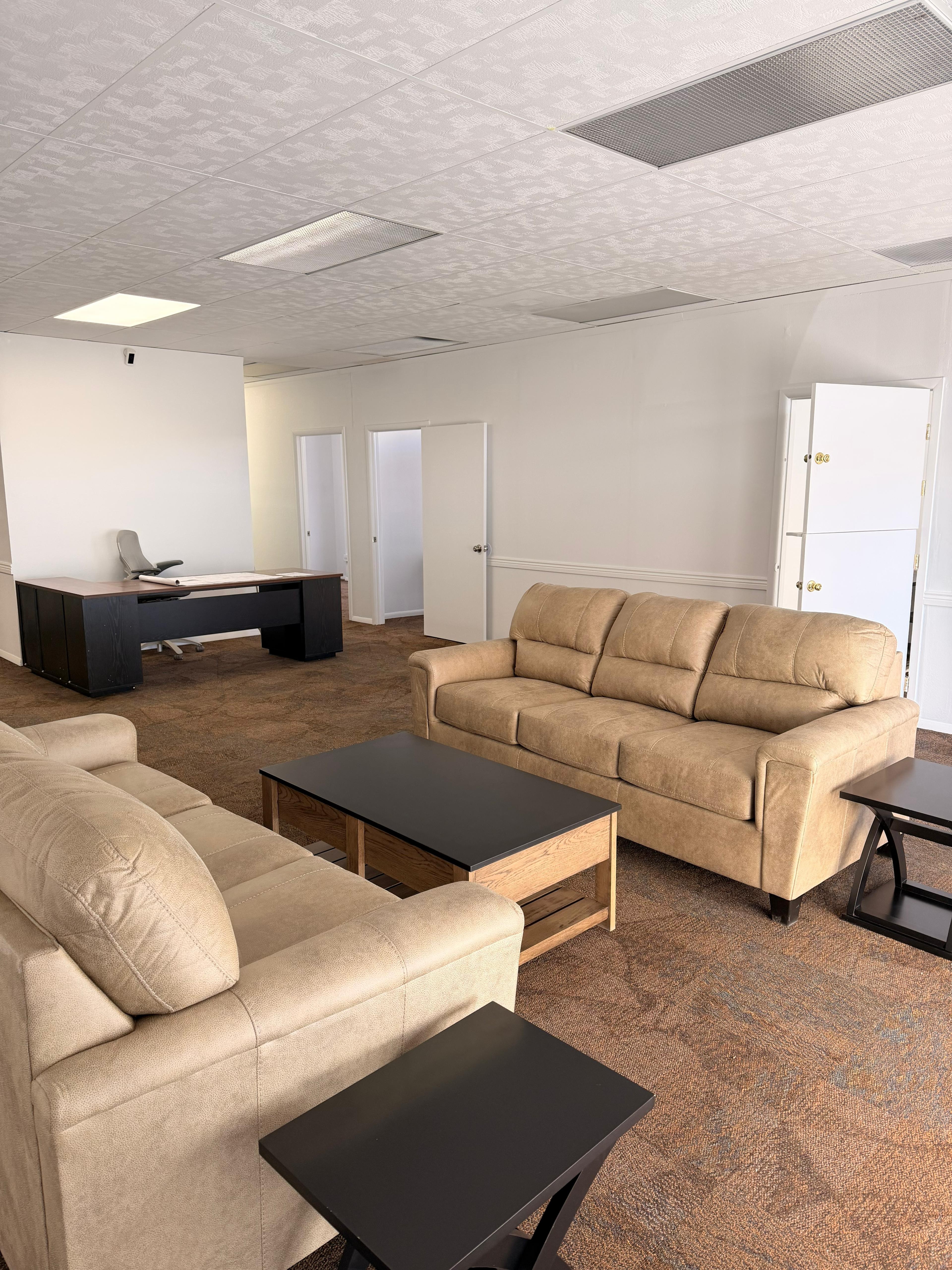 Waiting room with beige sofas, black coffee table, and white walls. Doors are open, ceiling has rectangular lights.