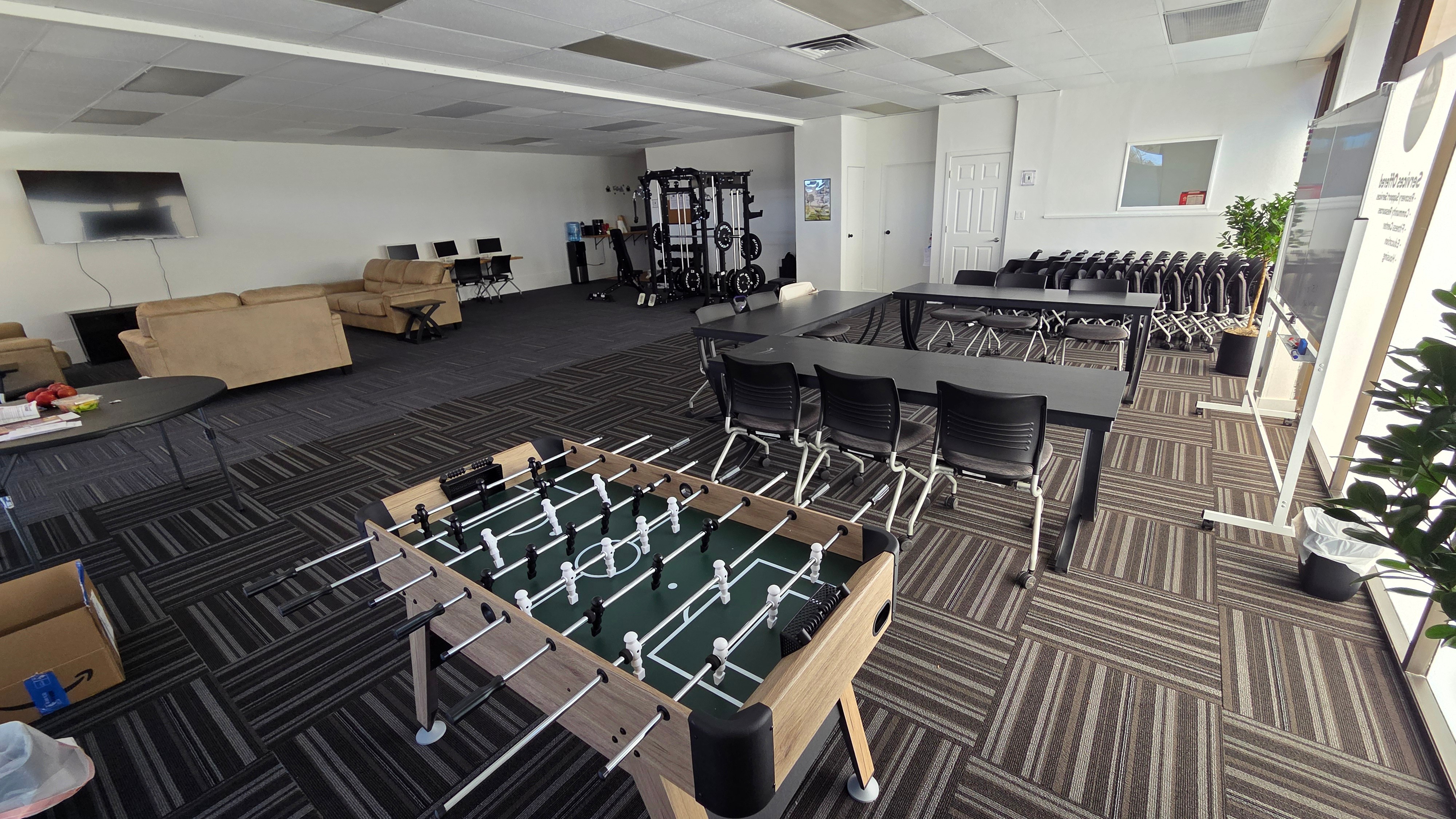 Large room with tables, chairs, a foosball table, and a lounge area, with windows and a TV on the wall.