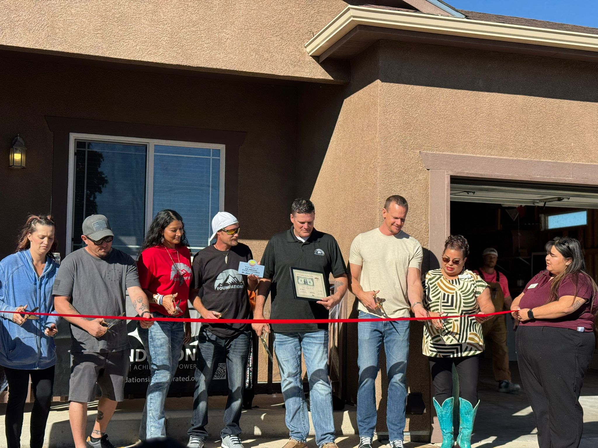 Group of people standing outside a house, holding a red ribbon for a celebration or event.