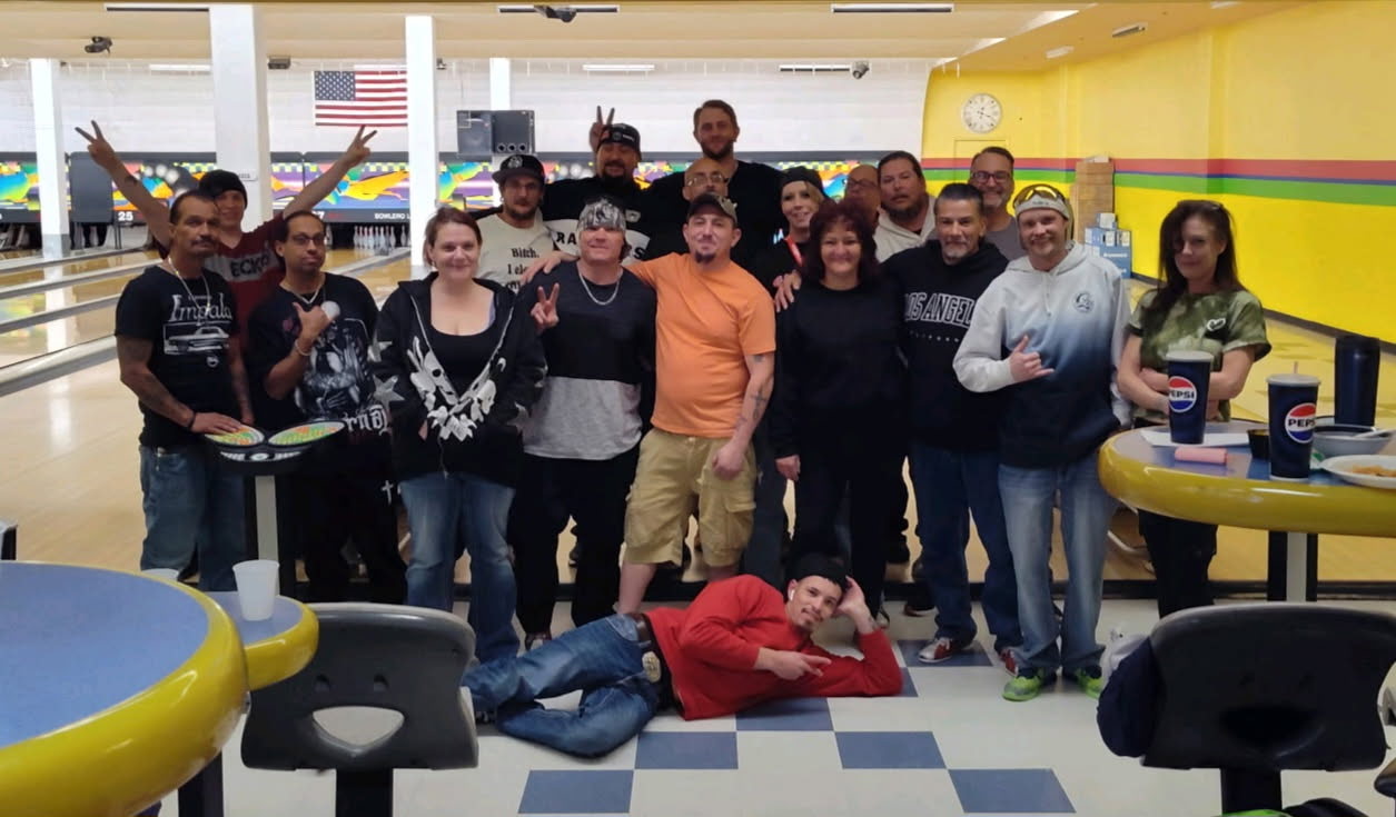 Group of people at a bowling alley, some standing and one lying on the floor, with bowling lanes in the background.