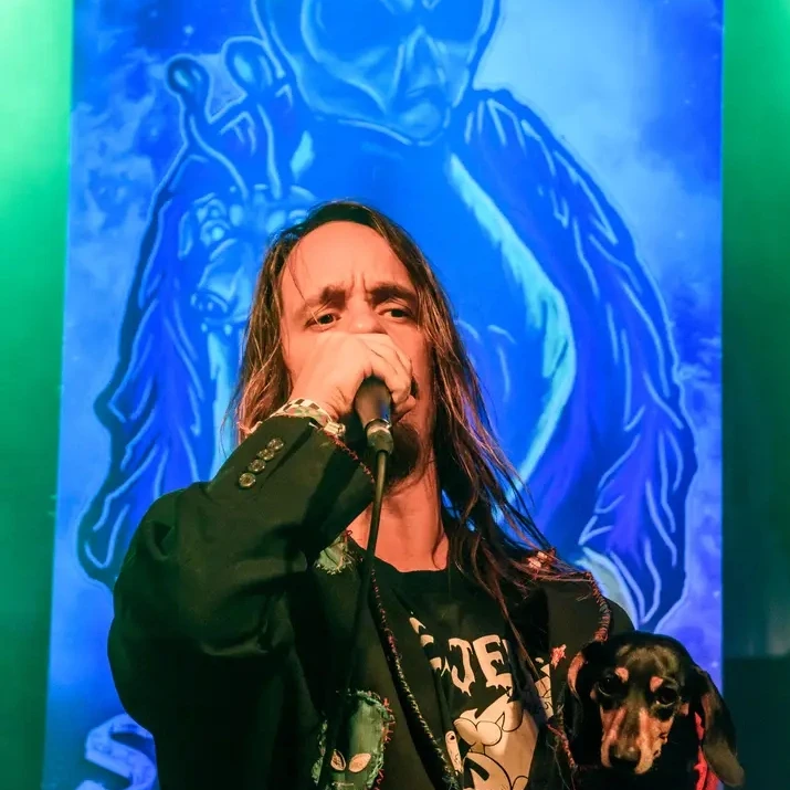 Person with long hair singing into a microphone, with a dog on their lap, in front of a blue background with a figure holding a sword.