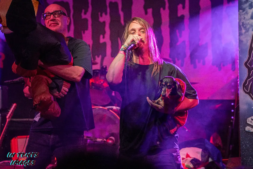 Two women on stage, one holding a child, with purple lighting and a backdrop, performing or speaking into a microphone.