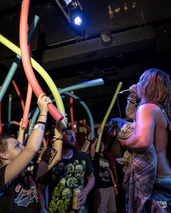 People holding colorful pool noodles in a dark indoor setting with a person speaking or singing.