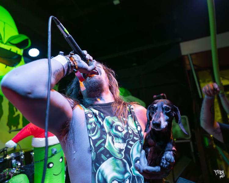 Person singing into a microphone while holding a small dog, with colorful stage background and green decorations.