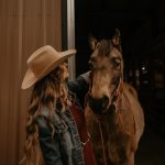 Person with long hair and wide-brimmed hat standing next to a horse inside a barn at night.