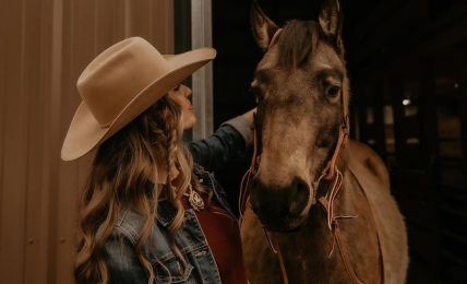 Person with long hair and wide-brimmed hat standing next to a horse inside a barn at night.