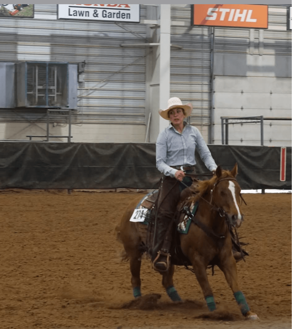 Person riding a horse in an indoor arena, wearing a hat and light-colored shirt, with banners overhead.