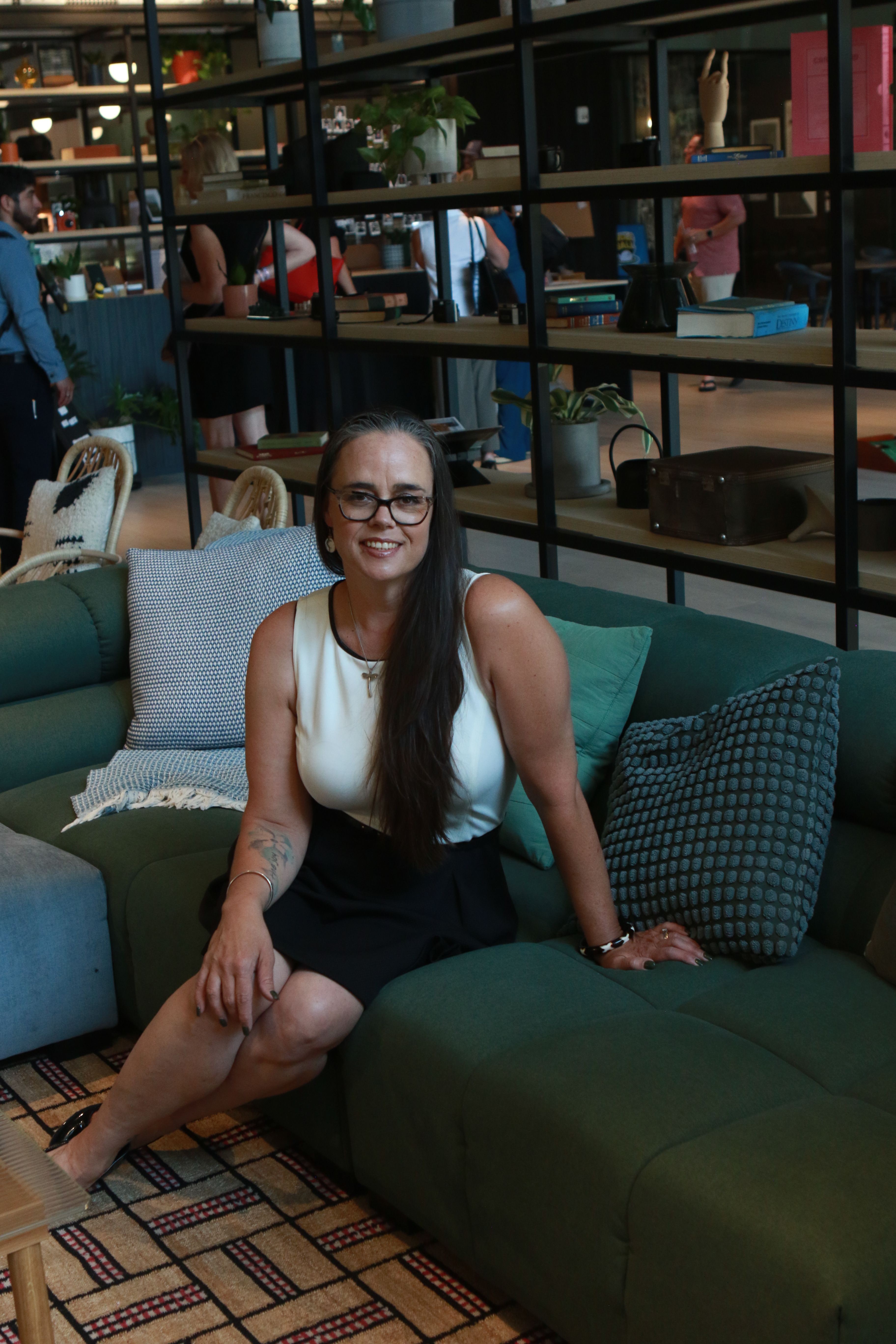 Woman with glasses and long hair sitting on a green sofa in a store, smiling, with shelves of products behind her.