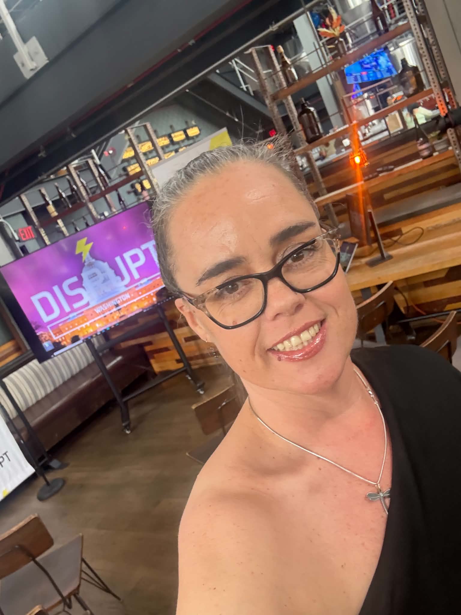 Woman with glasses smiling in a restaurant or bar setting, with a colorful sign and wooden furniture in the background.