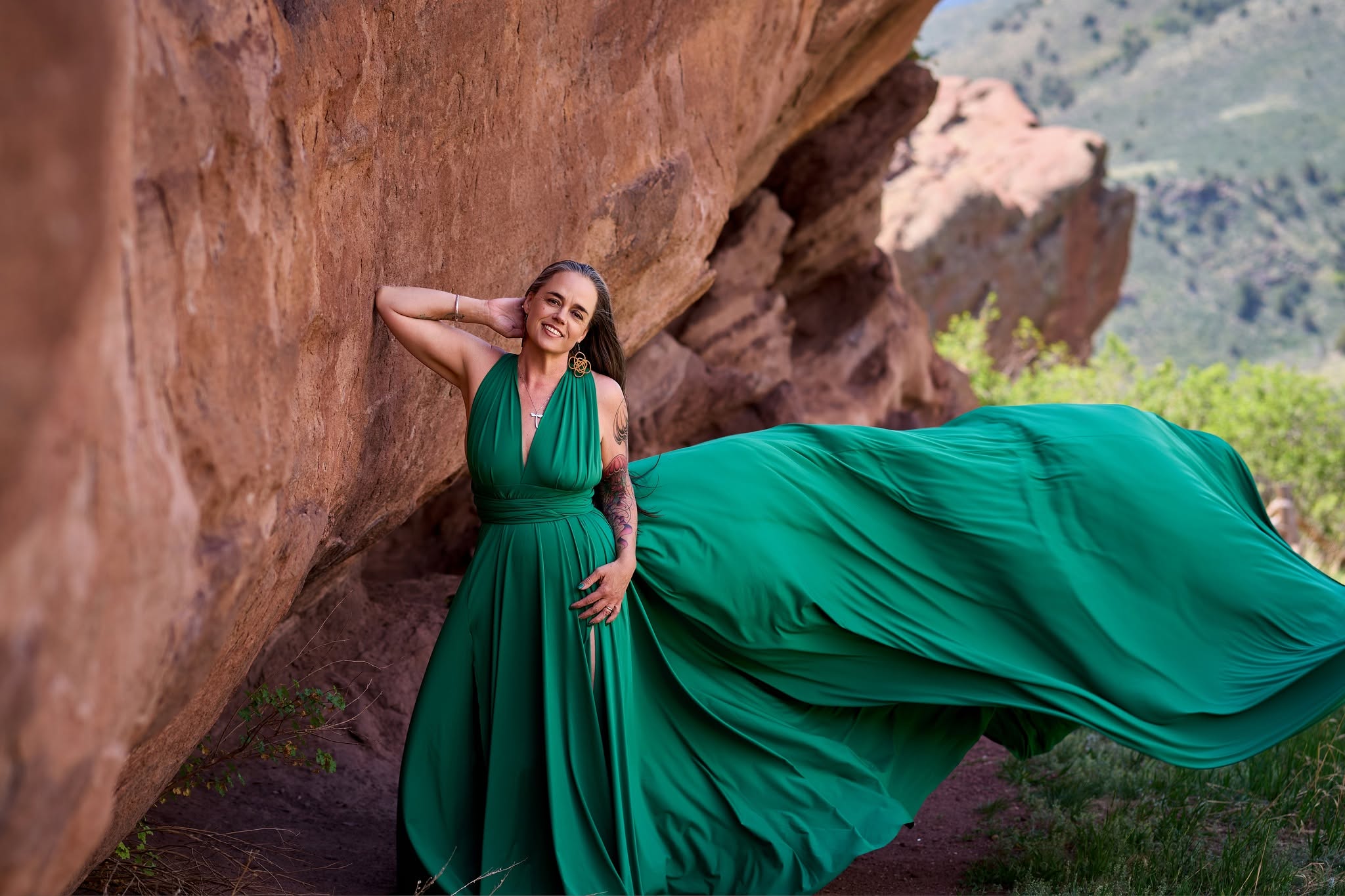 Woman in a flowing green dress leaning against a large rock outdoors, with mountains in the background.