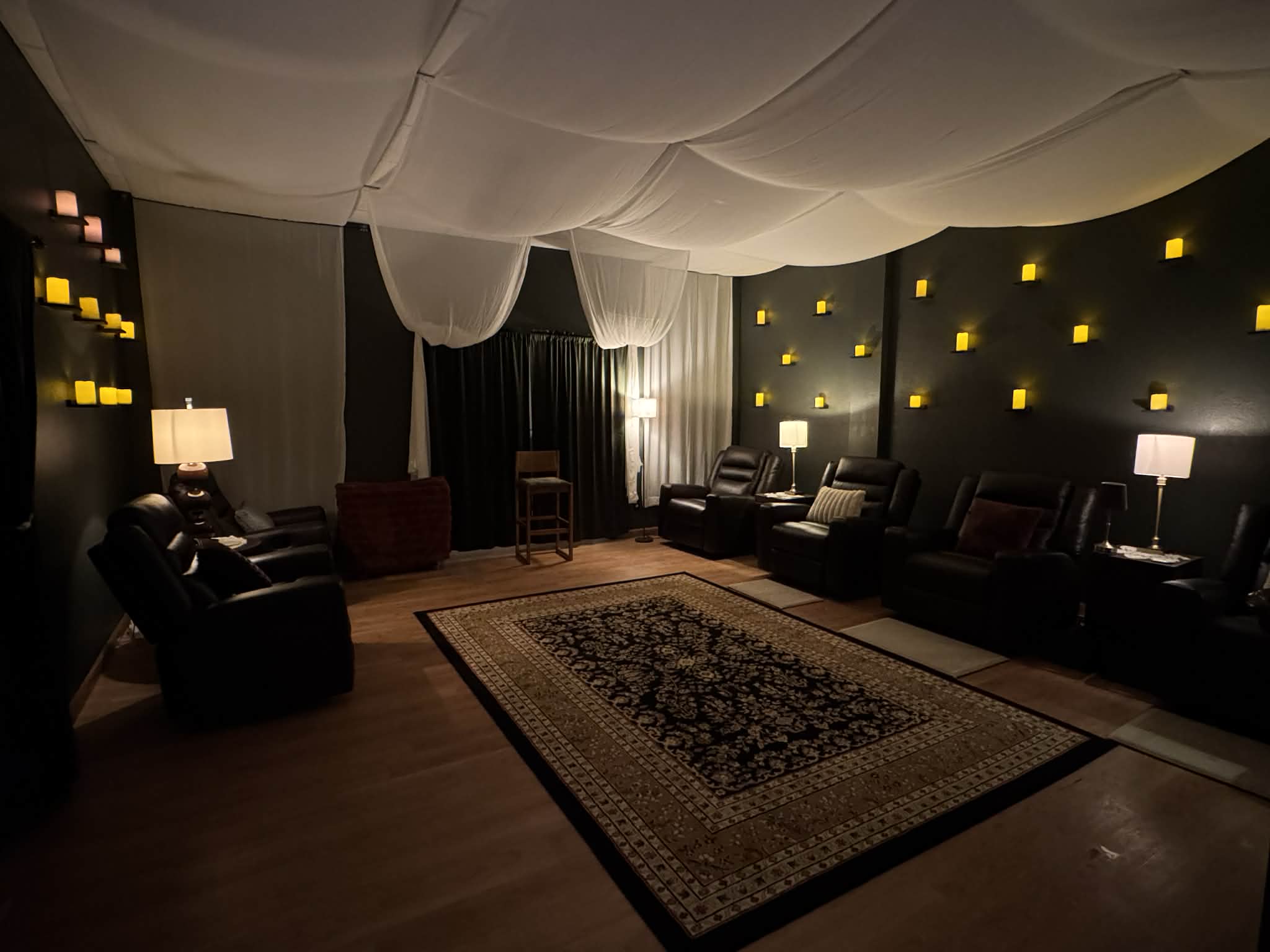 Room with dark walls, multiple armchairs, lamps, and a large patterned rug, ceiling draped with fabric, warm lighting.